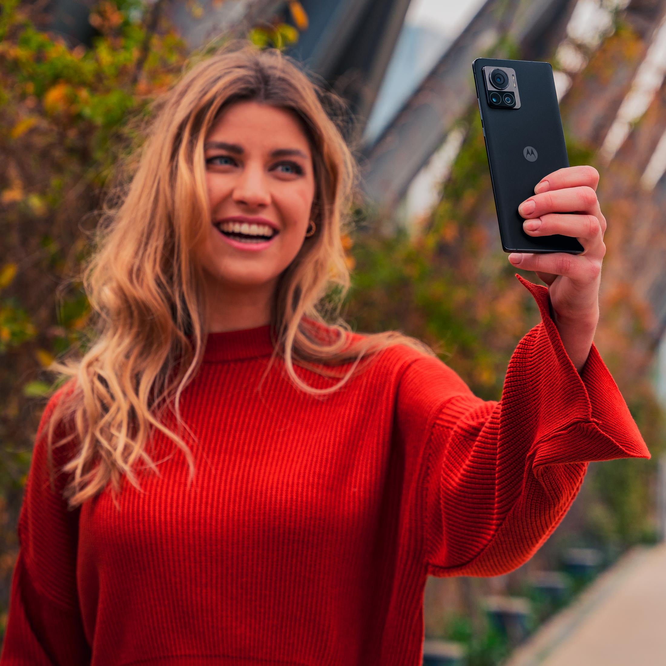 Frau macht ein Selfie mit einem Handy. Sie lächelt. Roter Pullover. Verschwommener Hintergrund.