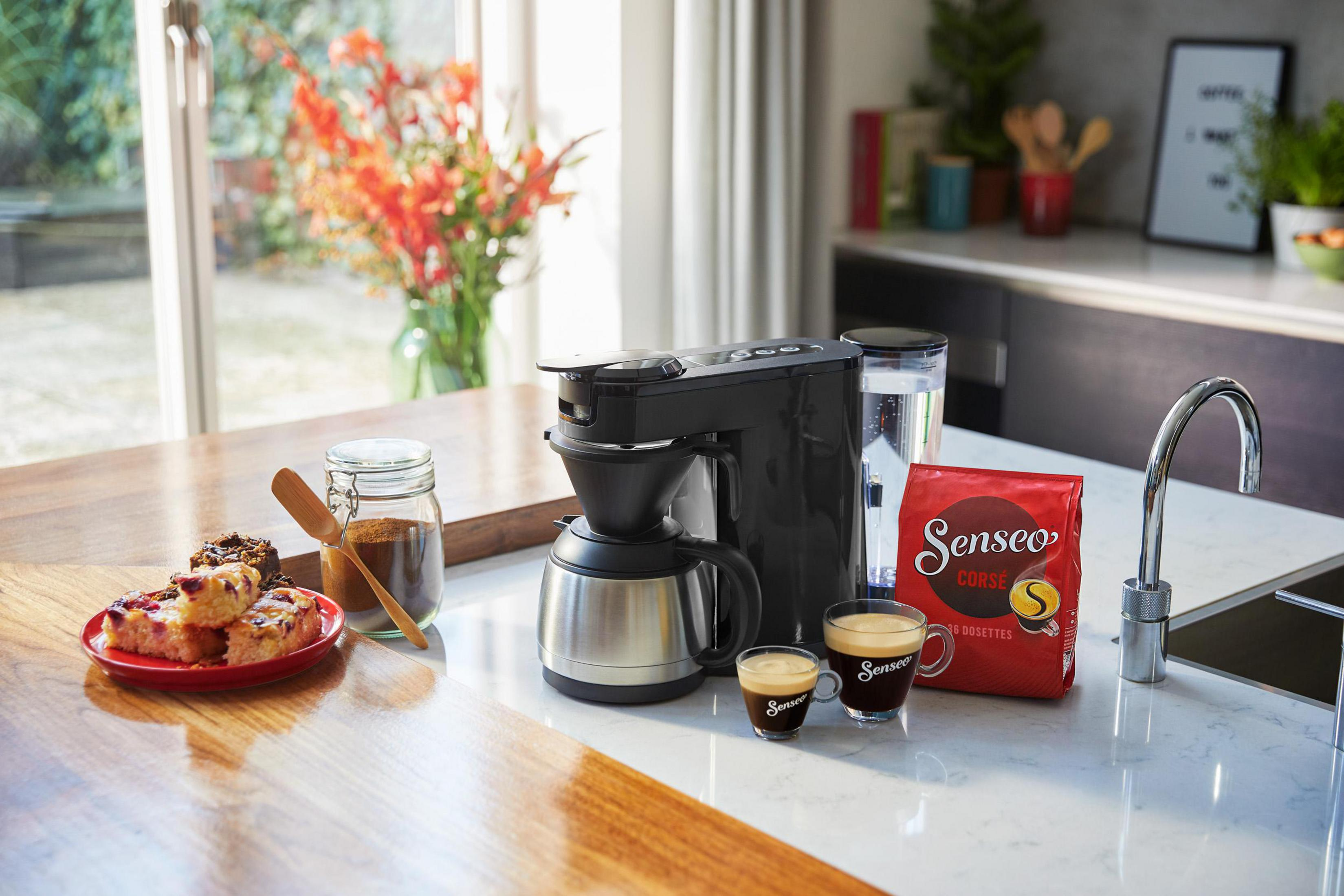 Ekspres do kawy, filiżanki i worek z ziarnami kawy. Jest stół z jedzeniem i oknem.