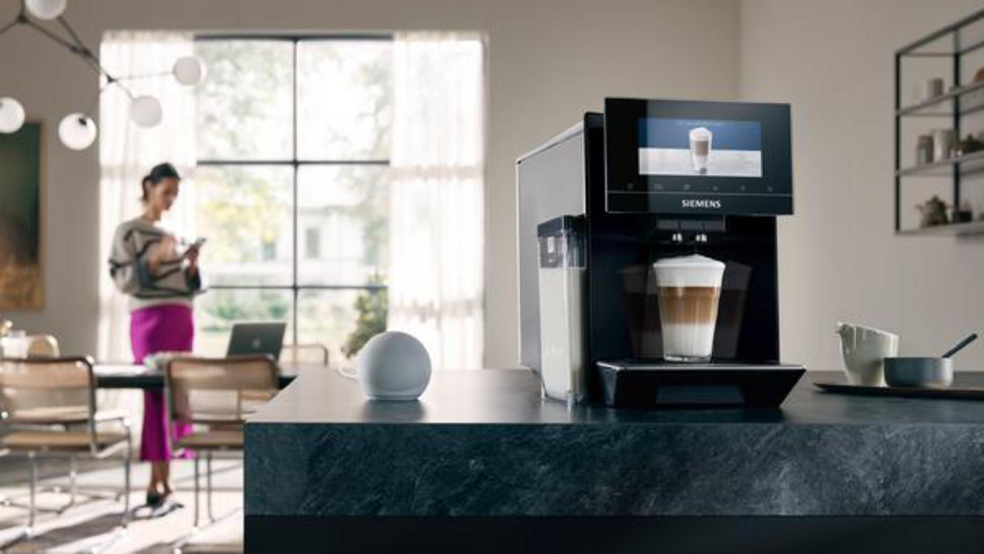 Una macchina da caffè che prepara il caffè con una donna sullo sfondo. È presente anche un altoparlante intelligente.