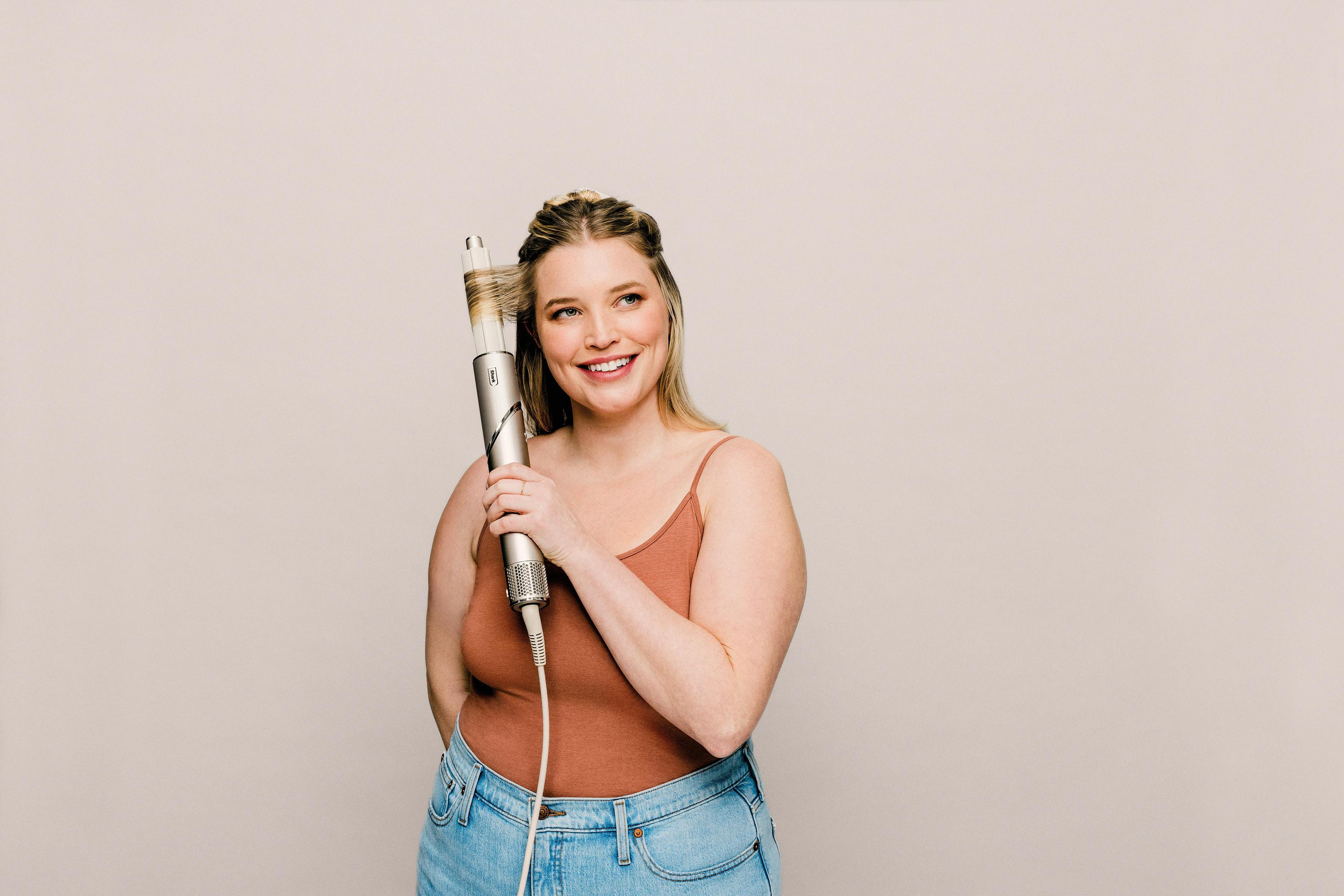 Kobieta trzymająca lokówkę, uśmiechnięta. Beżowe tło.