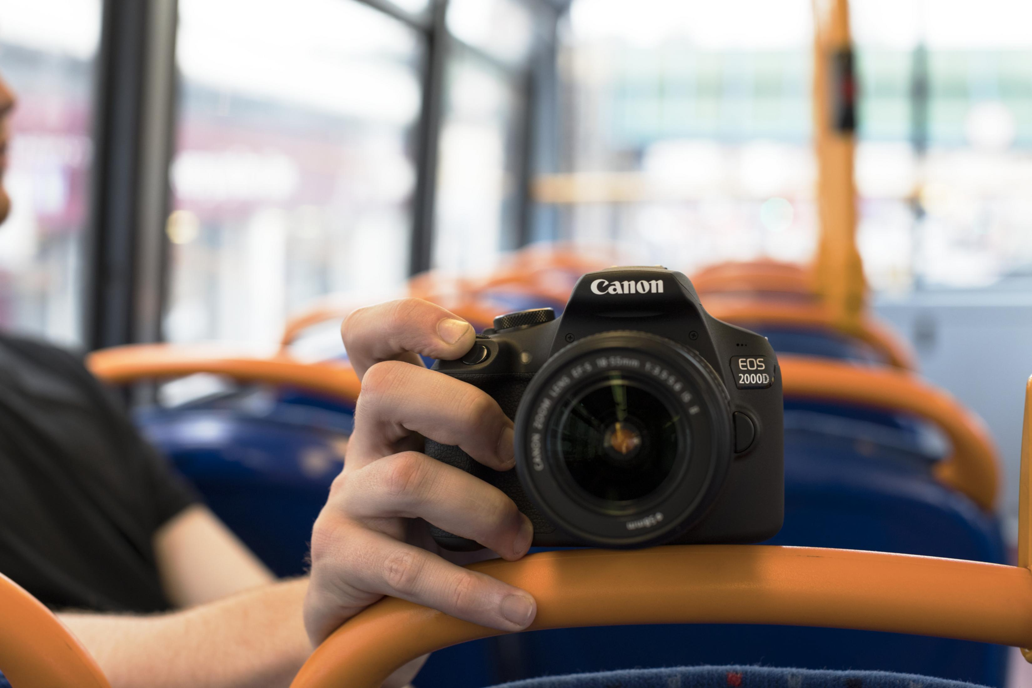 Osoba trzymająca aparat Canon w autobusie, skupiając się na obiektywie, z siedzeniami autobusu w tle.