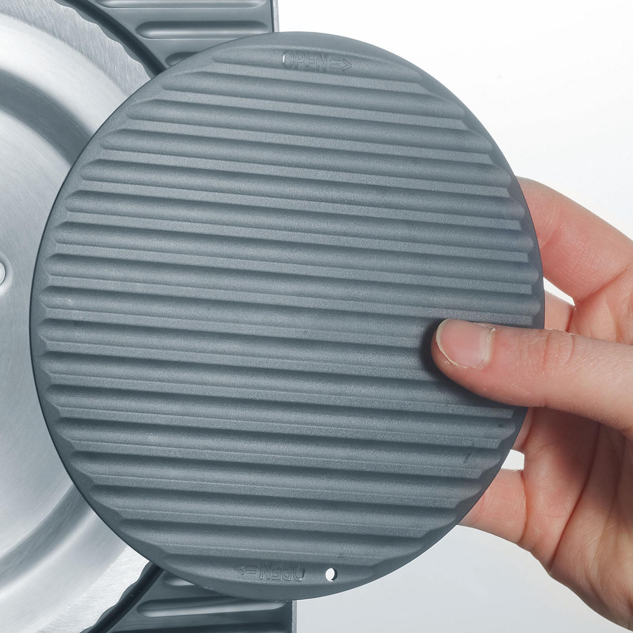 Szara okrągła pokrywka z rowkami, trzymana przez dłoń, w pobliżu metalowej powierzchni.