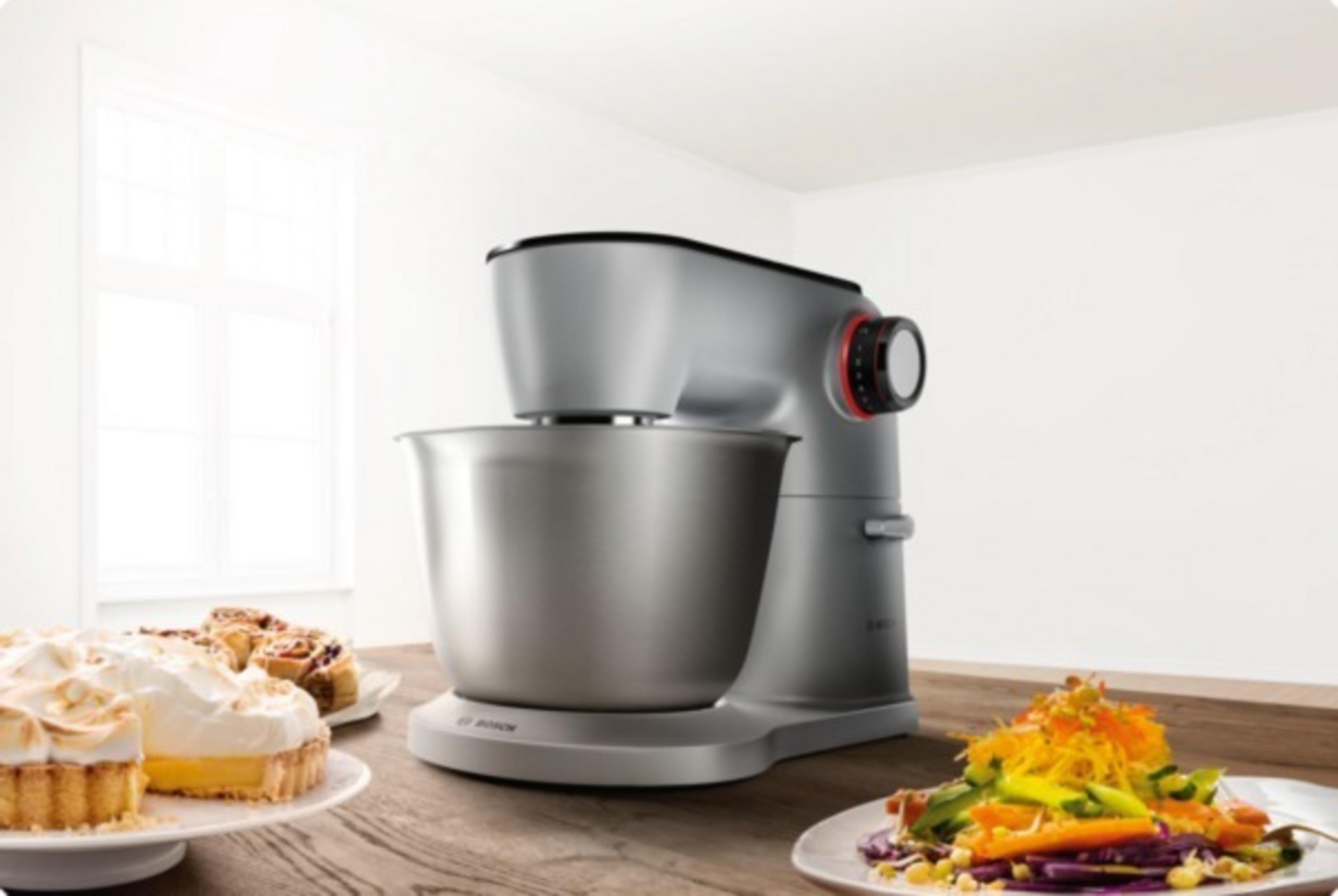 Ein silberner Küchenmixer mit Essen auf Tellern. Kuchen, Torten und Salat auf dem Holztisch.