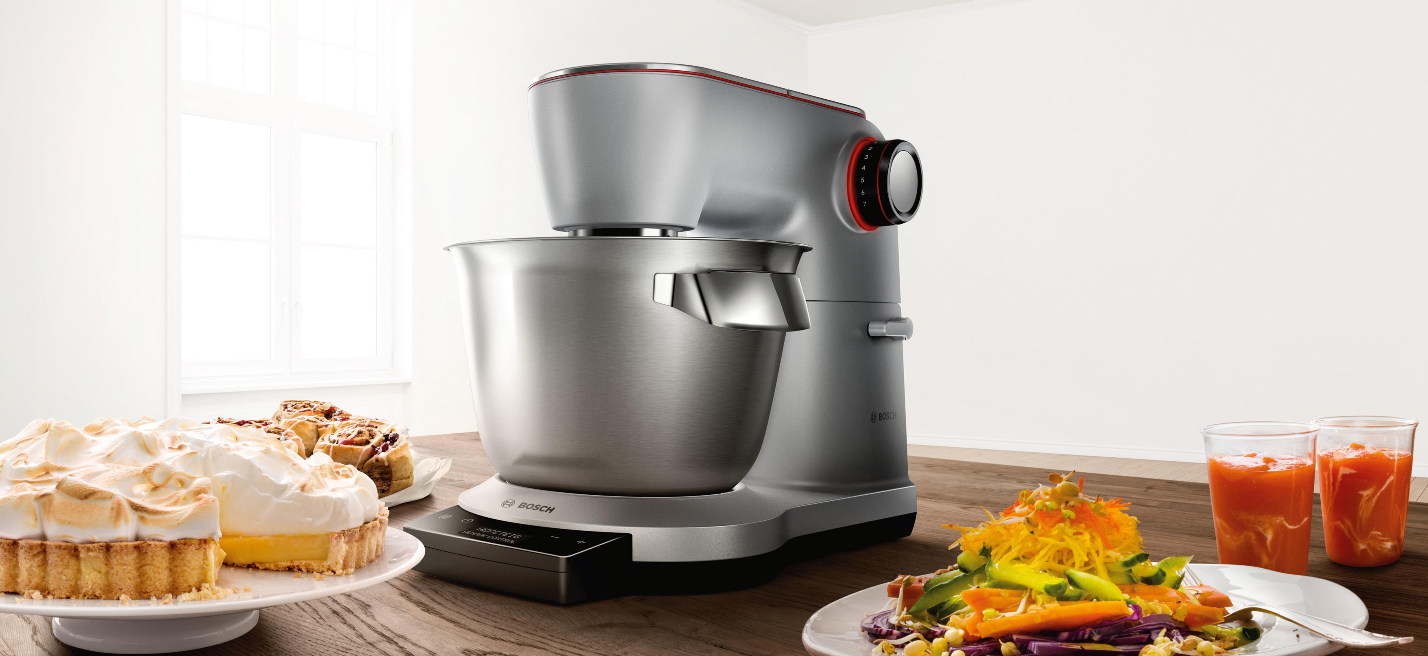 Silberfarbener Küchenmixer mit Essen, Desserts auf Holztisch, weißer Wandhintergrund.