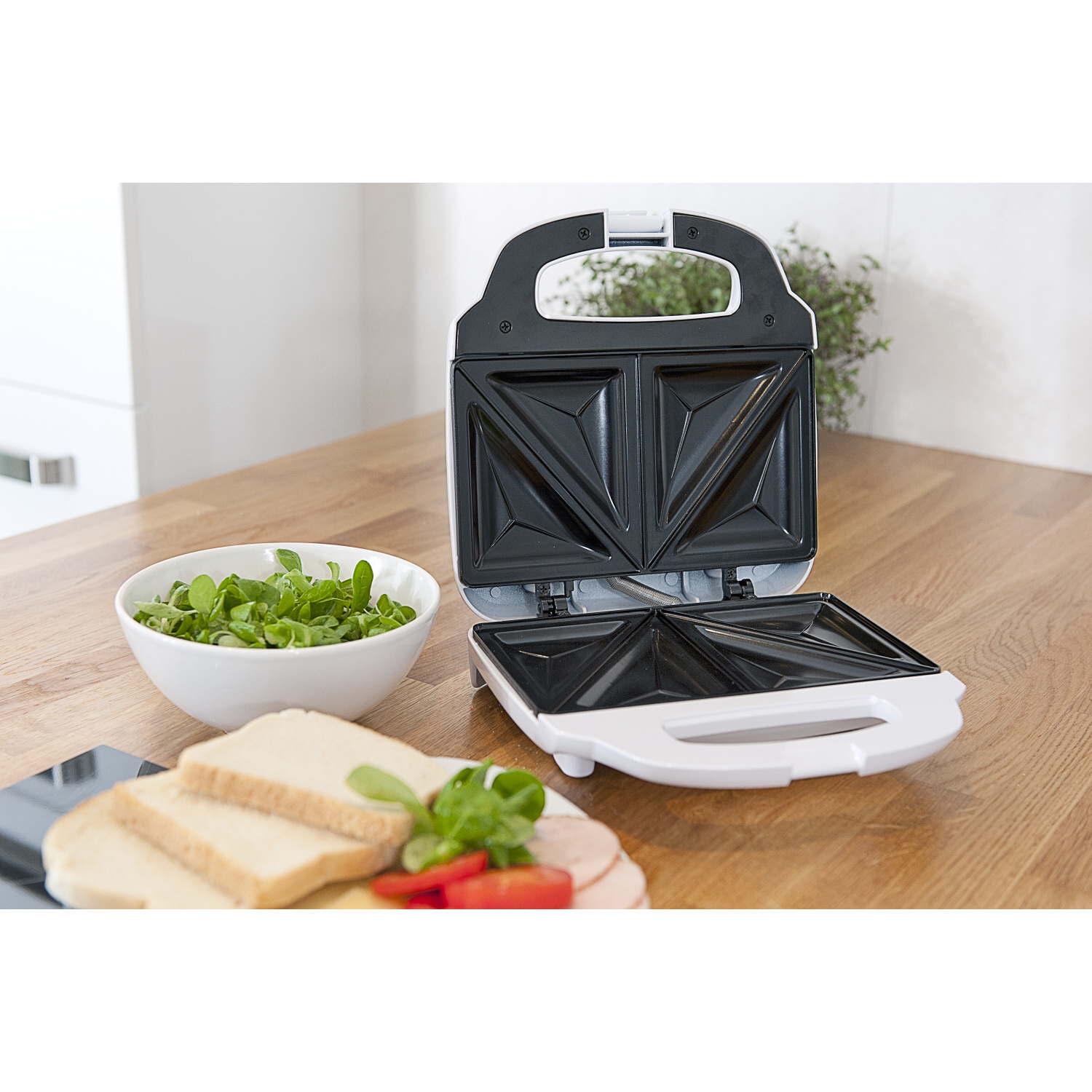 Sandwichera abierta con mitades de sándwich, un cuenco de verduras y rebanadas de pan sobre una mesa de madera. El aparato es blanco.