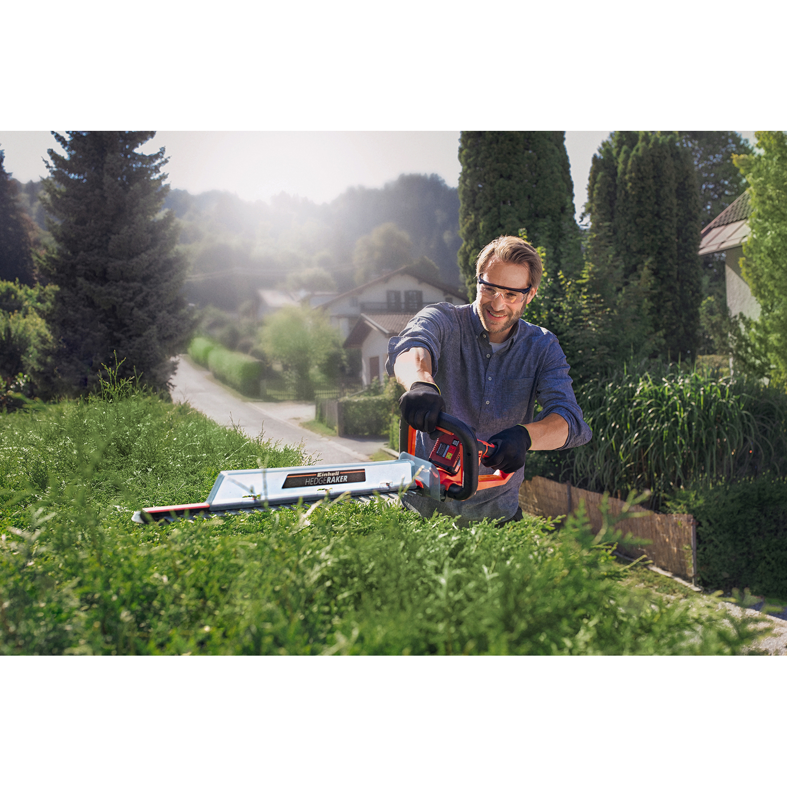 Ein Mann mit Brille und Handschuhen benutzt eine Heckenschere an einer grünen Hecke im Freien bei Sonnenschein.