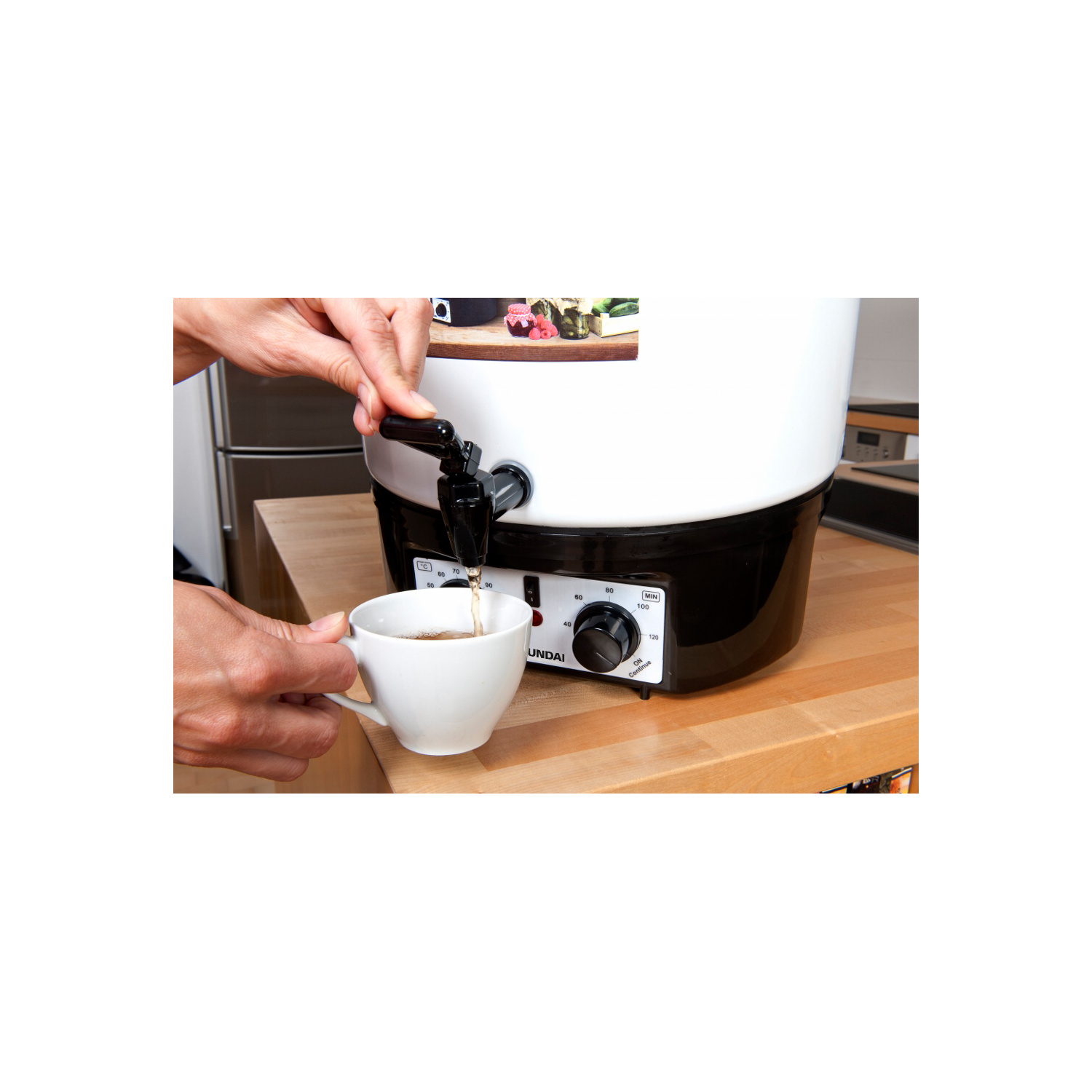 Eine Person gießt Tee aus einem weiß-schwarzen Spender in eine weiße Tasse auf einer Holztheke.