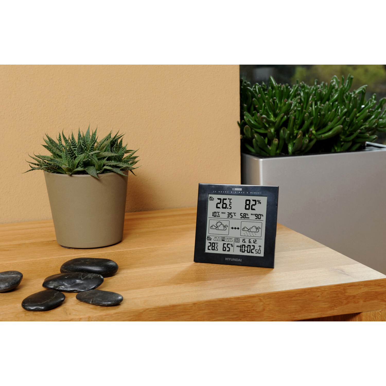 Ein Holztisch mit einer Digitaluhr und einigen schwarzen Steinen. Ein Blumentopf mit einer Pflanze und eine grüne Pflanze ist sichtbar.