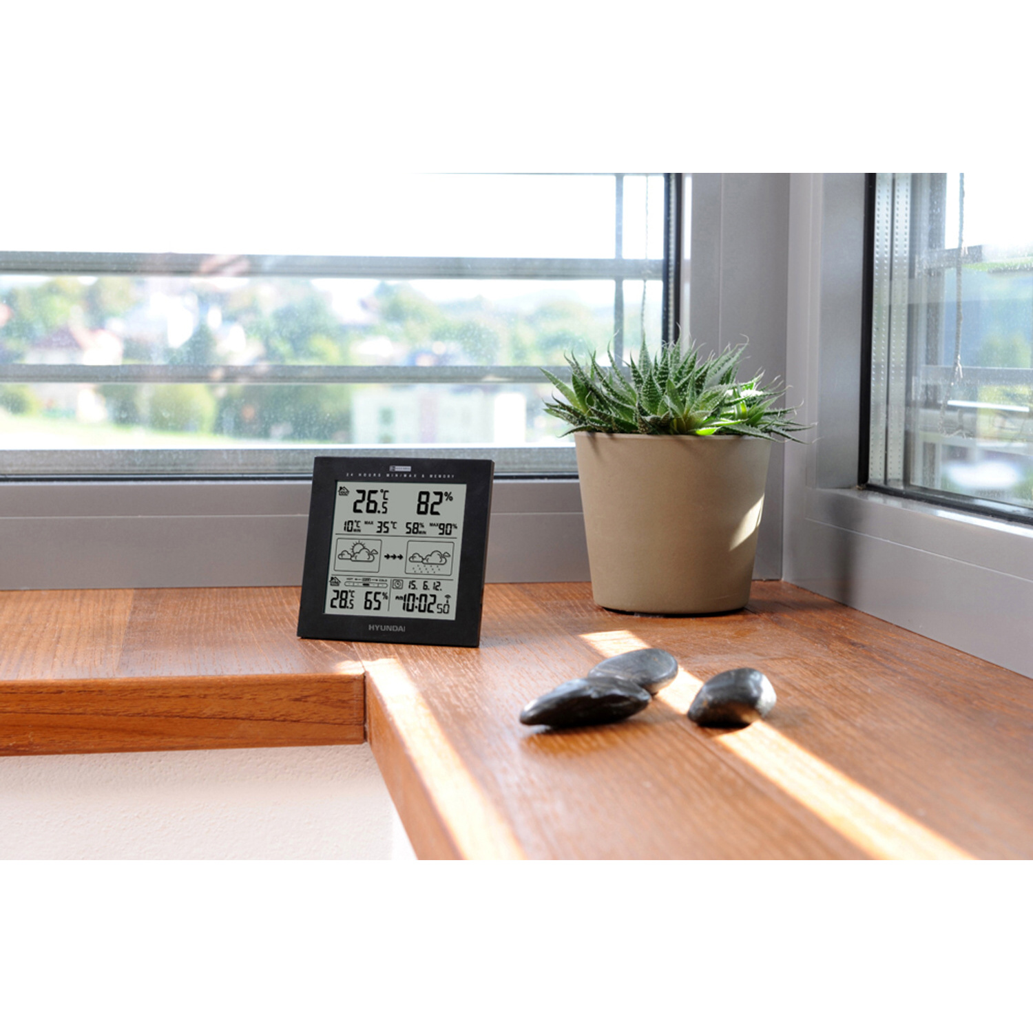 Schwarze digitale Wetterstation am Fenster, mit Pflanze und Steinen.