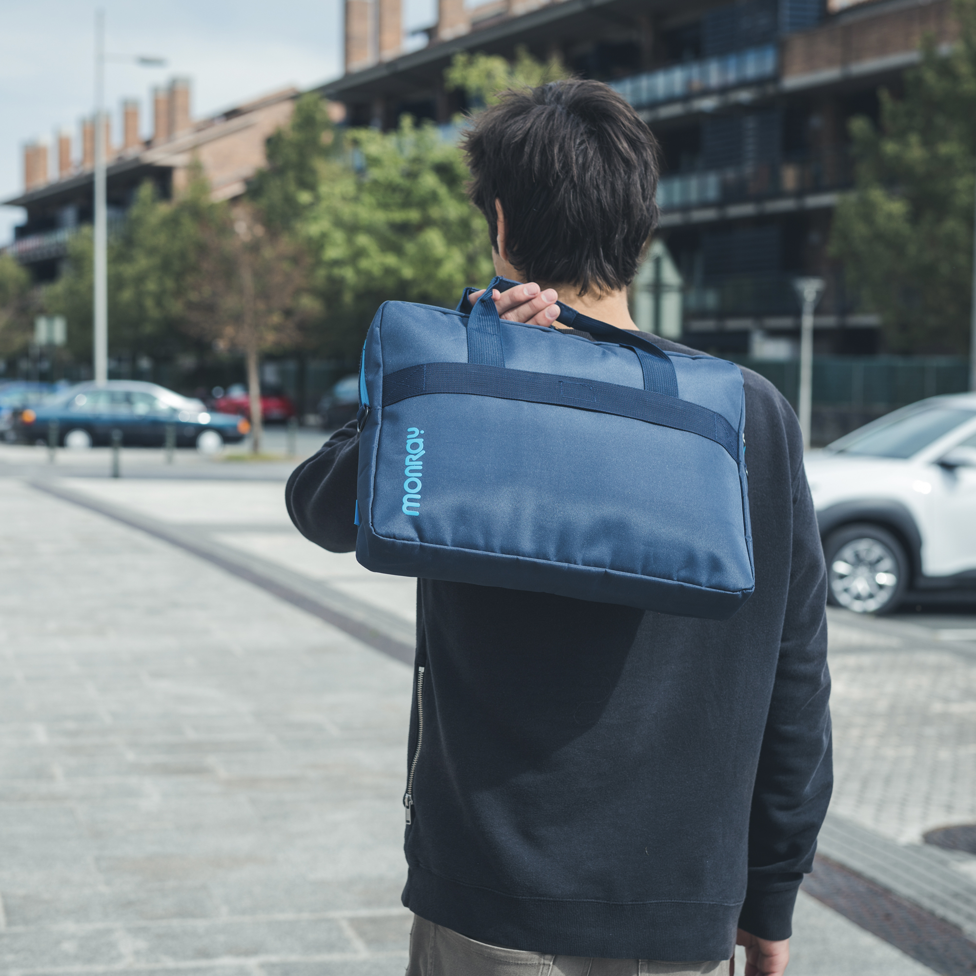 Ein Mann geht auf einem Bürgersteig und trägt eine blaue Tasche mit dem Wort MONROY darauf.