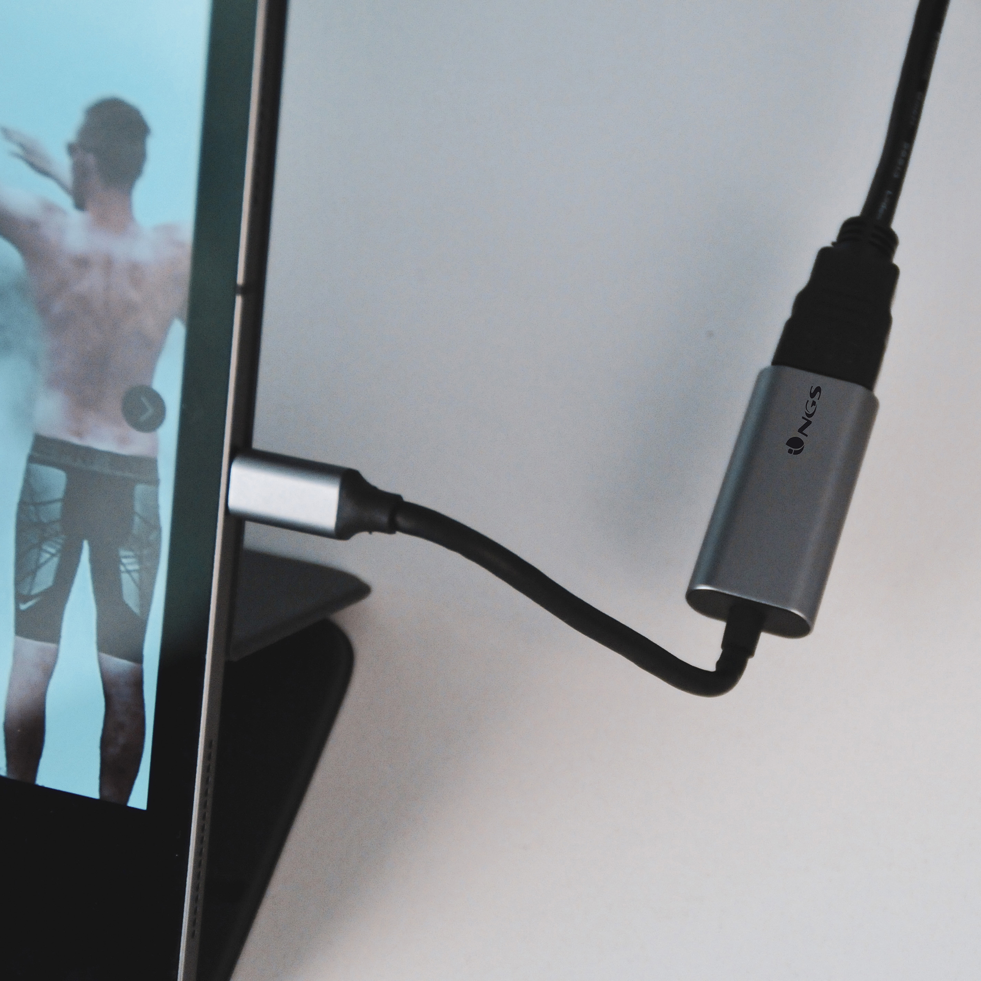 Una tableta está conectada a un adaptador USB-C plateado. Un hombre con la espalda desnuda es visible en la pantalla.