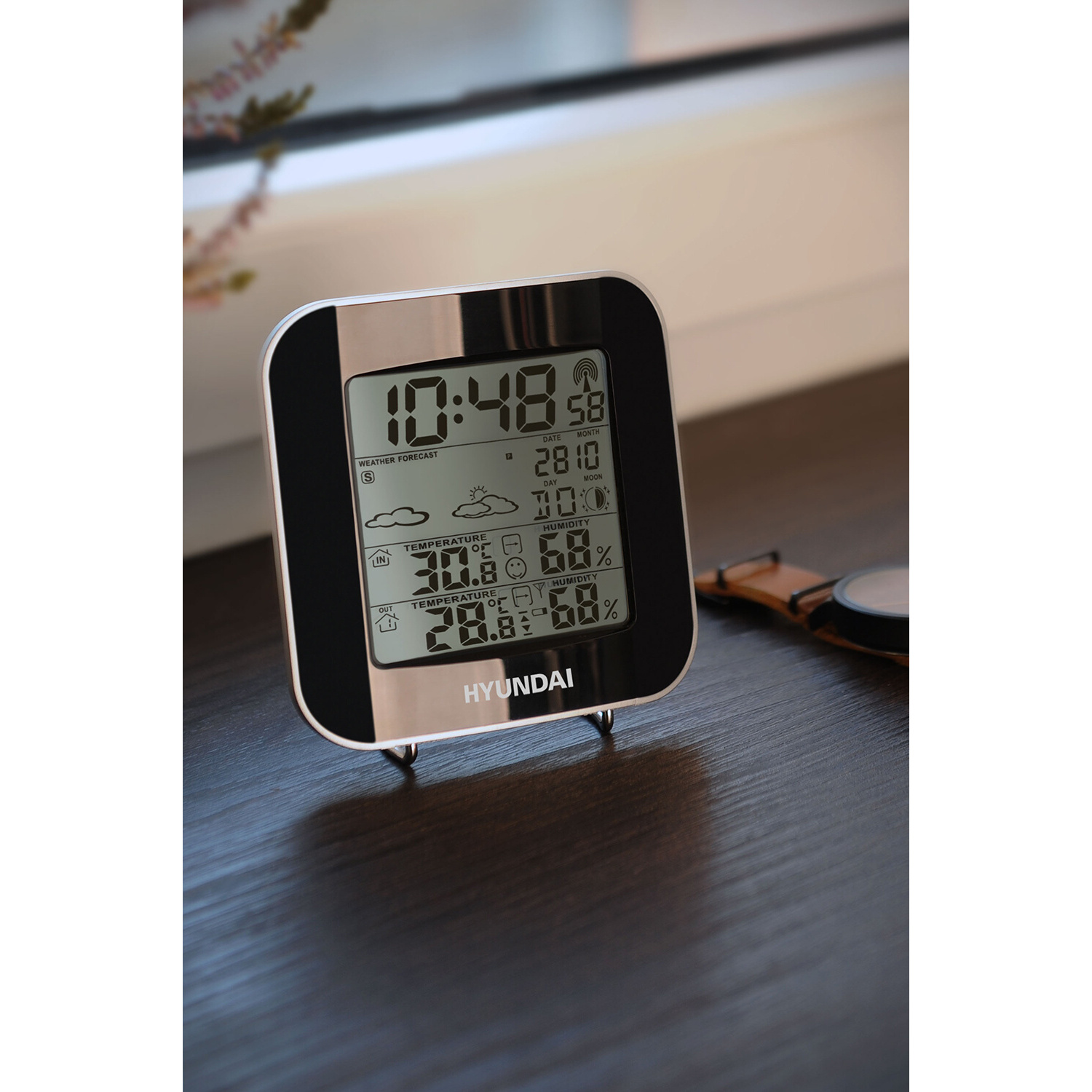 Digitaluhr mit Zeit-, Datums- und Wetterinformationen. Sie steht auf einem Tisch.