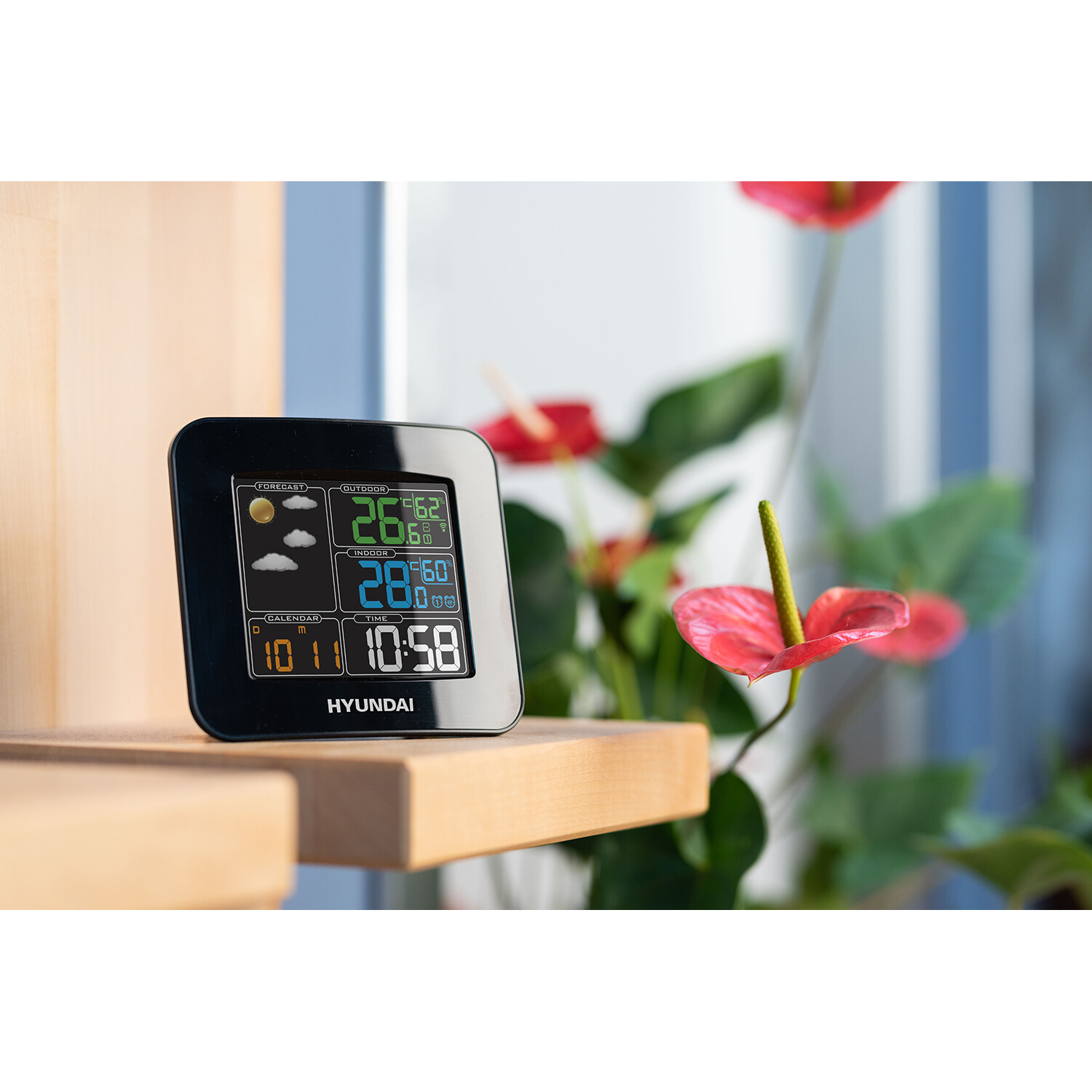 Schwarze Wetterstation, die Wetterdaten anzeigt. Rote Blumen und Holzoberfläche im Hintergrund.