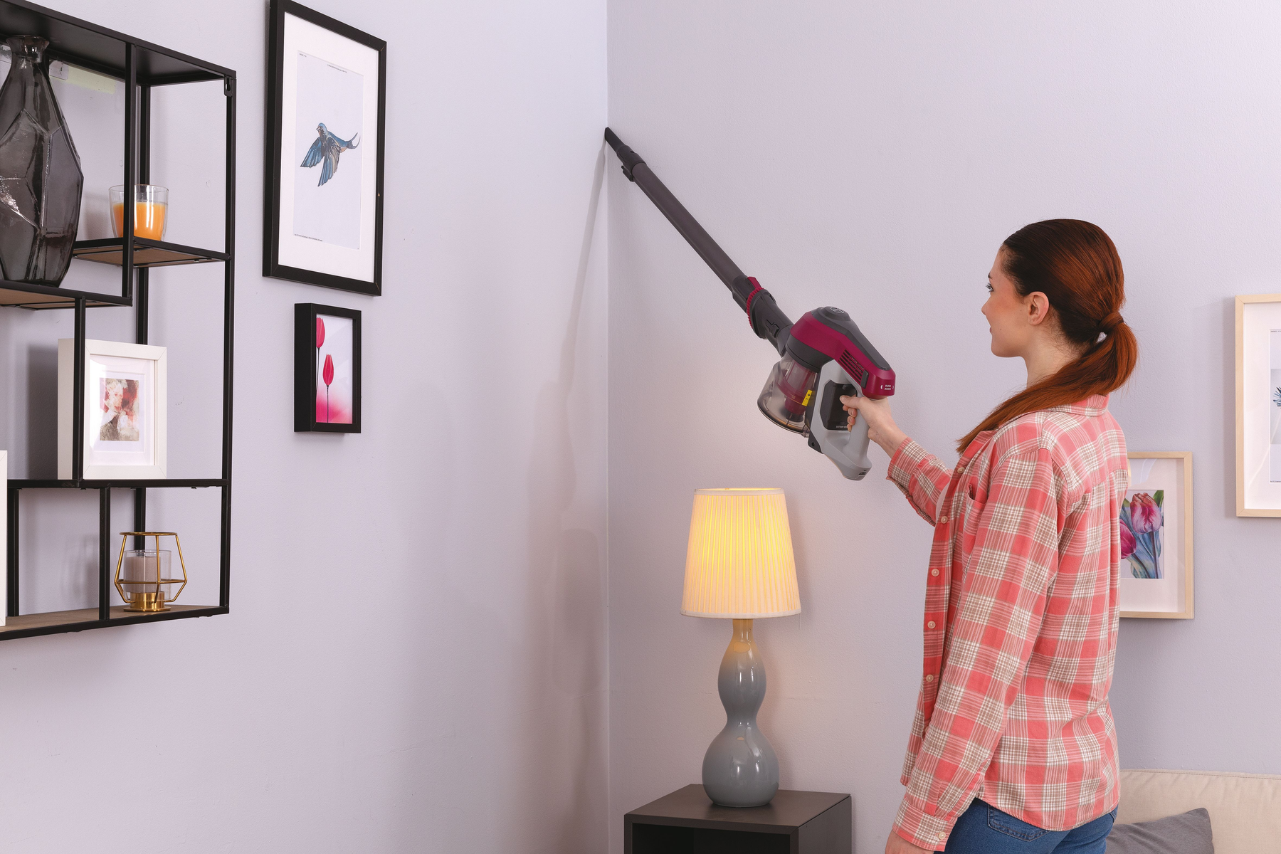 Eine Frau saugt eine Wand-Ecke mit einem Handstaubsauger, in der Nähe von Regalen und Lampen.