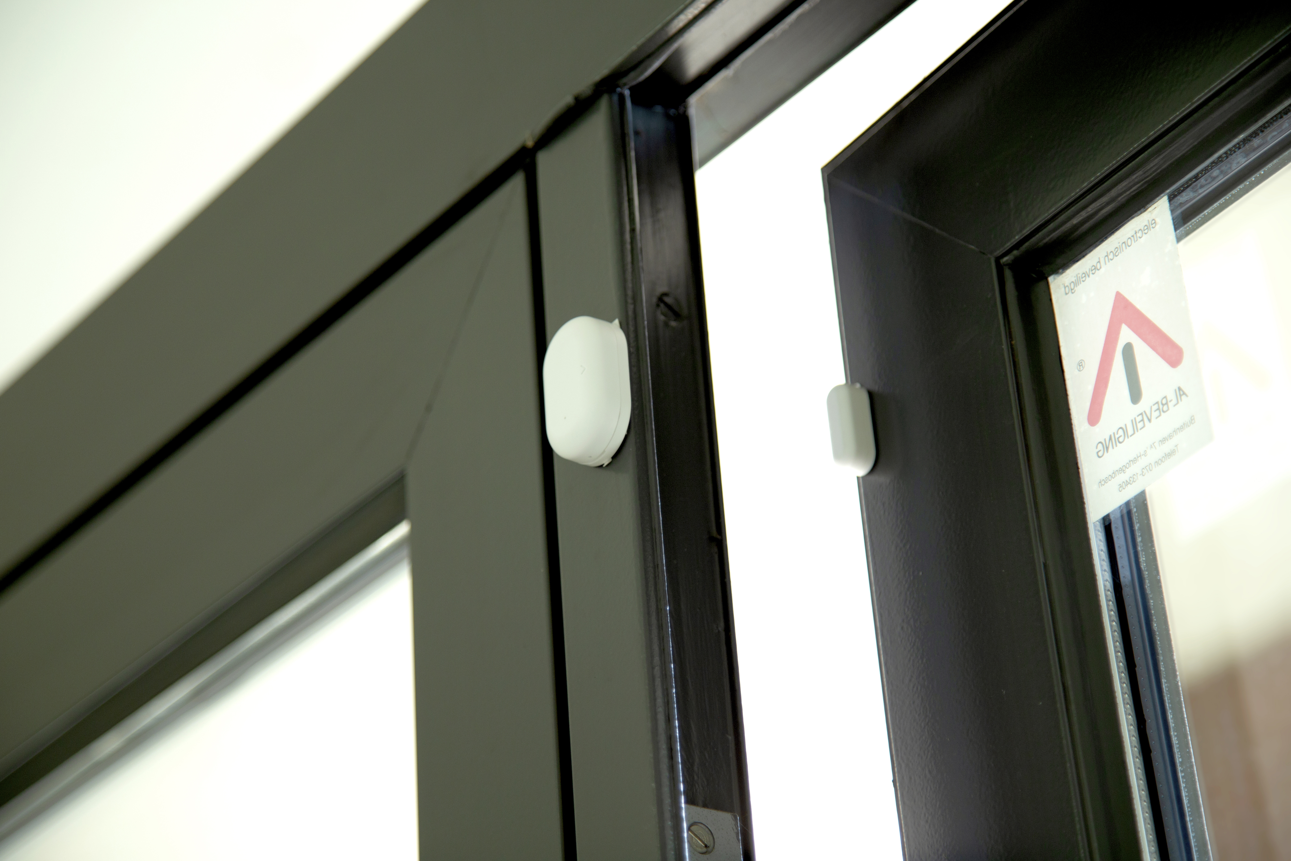 Fensterrahmen mit Sicherheitssensor. Ein weißer Sensor ist am Rahmen montiert. Text auf dem Glas sichtbar.