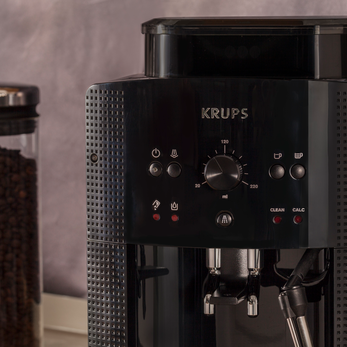 Cafetera Krups negra. Tiene un dial para ml, botones y un recipiente transparente con granos de café.