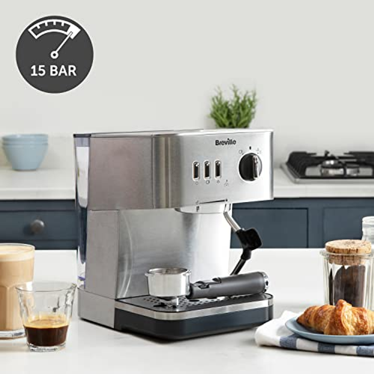 Máquina de espresso Breville de acero inoxidable con accesorios, una taza de café y un cruasán.