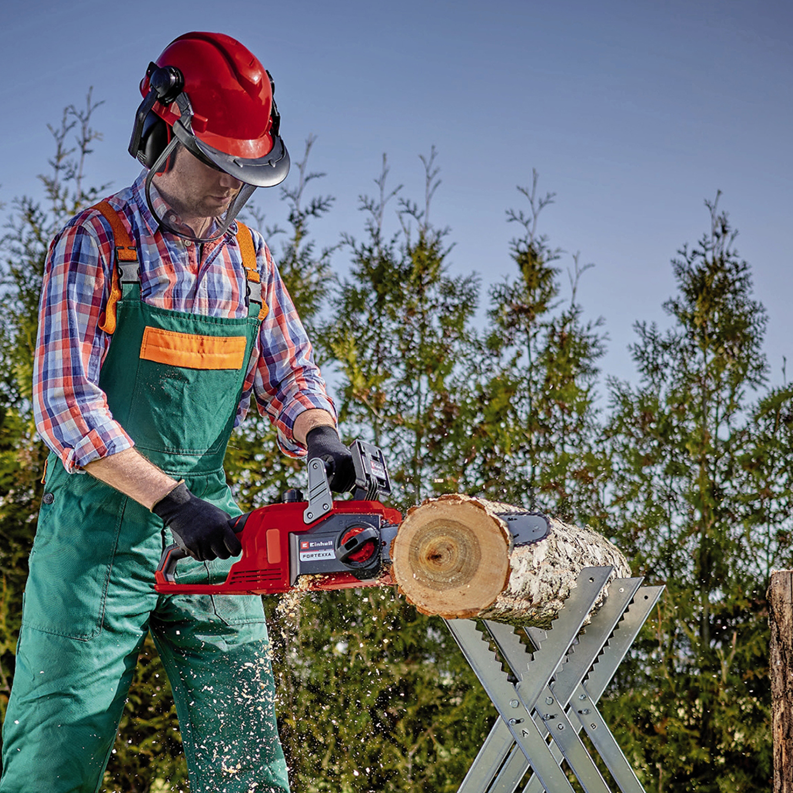 Mężczyzna używający piły łańcuchowej do cięcia kłody. Widoczne wyposażenie ochronne. Zielone drzewa w tle.