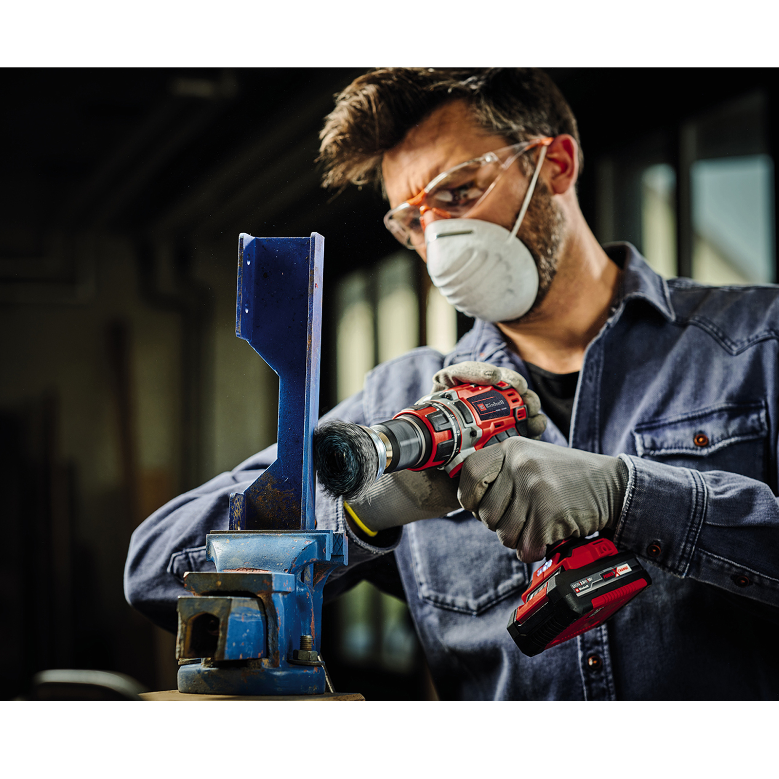 Ein Mann mit Maske und Brille verwendet eine Bohrmaschine an einem Metallobjekt, das in einem Schraubstock eingespannt ist.