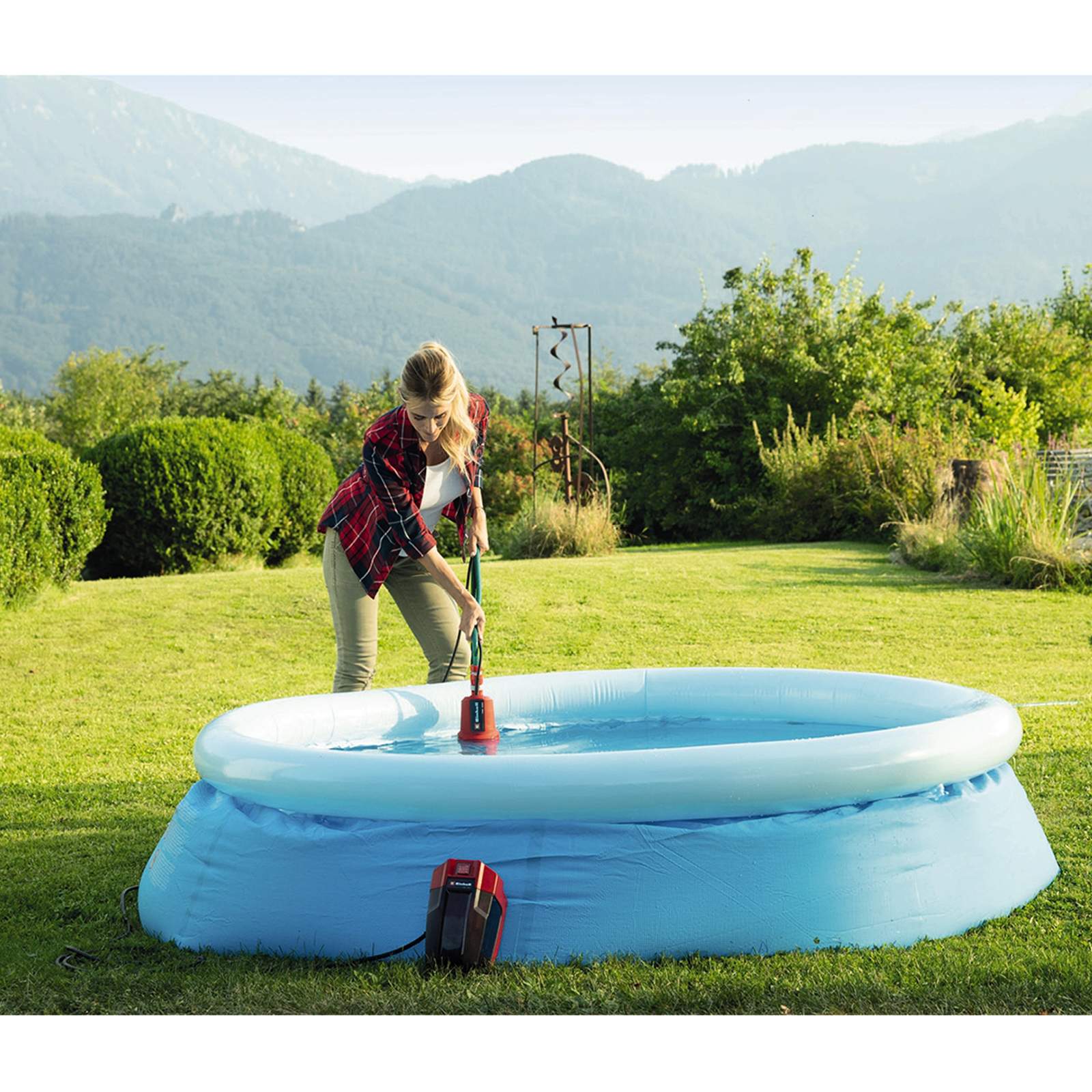 Kobieta napełnia niebieski basen dmuchany pompą w ogrodzie, w tle góry.