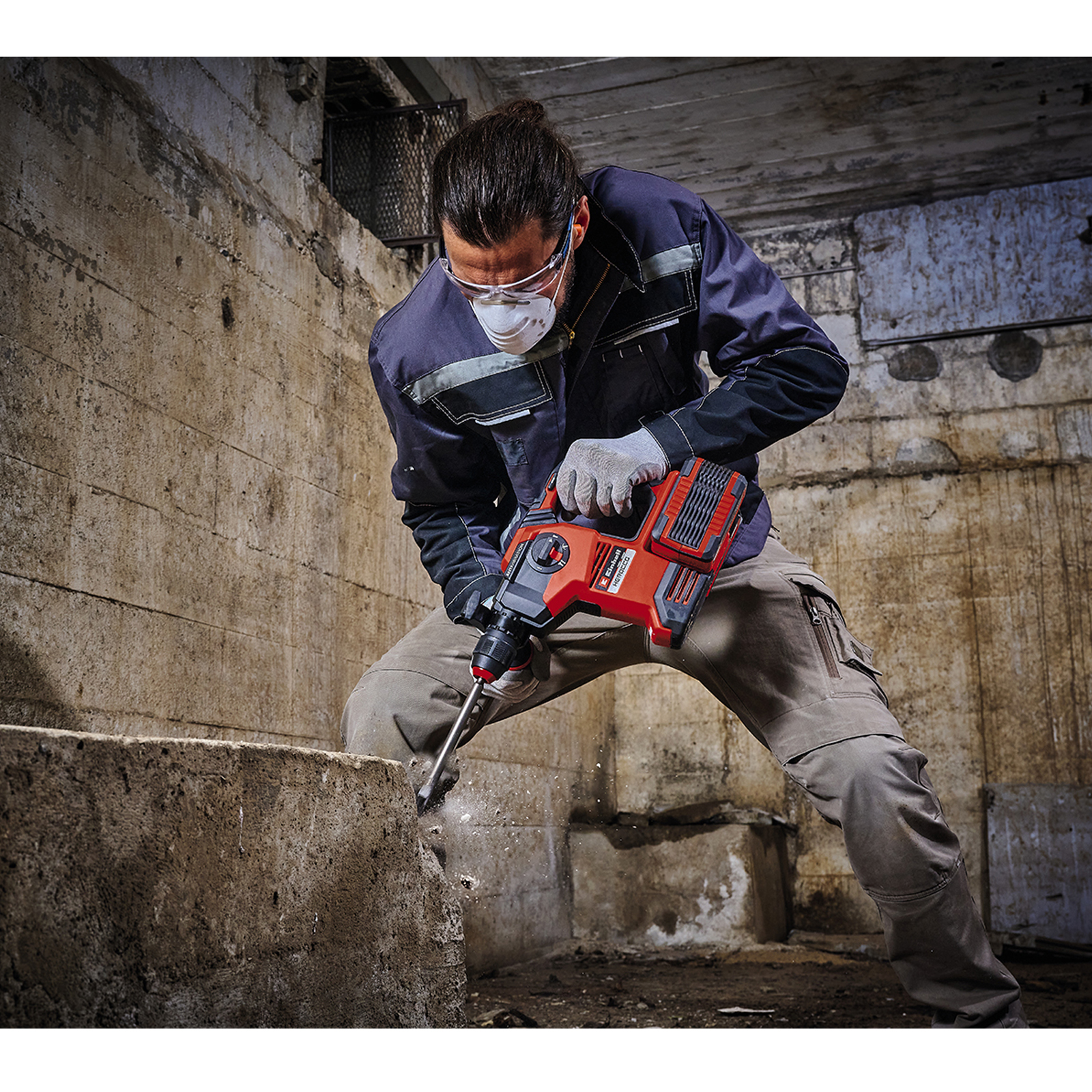 Mężczyzna w okularach ochronnych i masce wierci w betonie czerwoną wiertarką. Scena w ciemnym, betonowym pomieszczeniu.