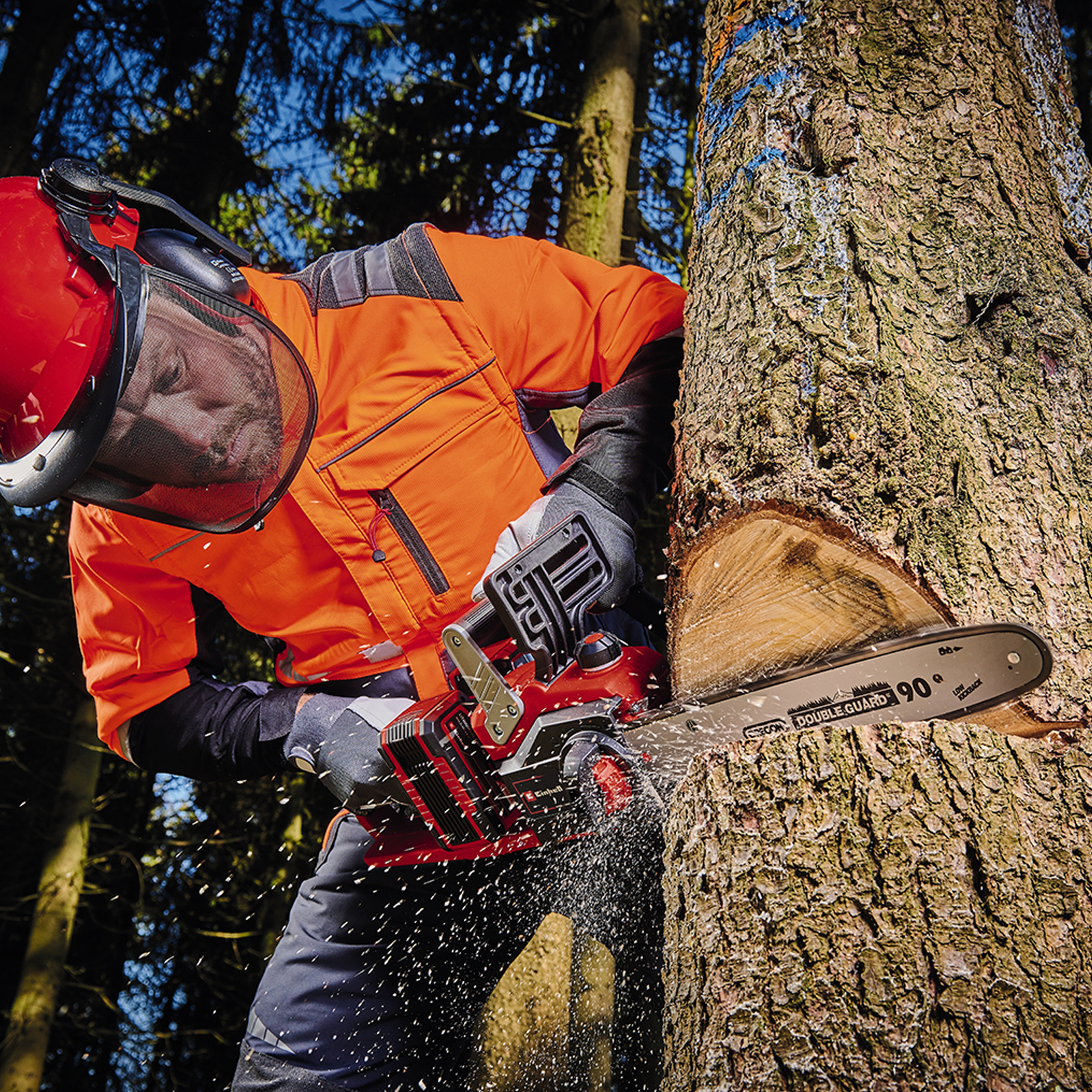 Mężczyzna w pomarańczowej kurtce używa piły łańcuchowej do cięcia pnia drzewa. Wióry lecą w powietrzu.