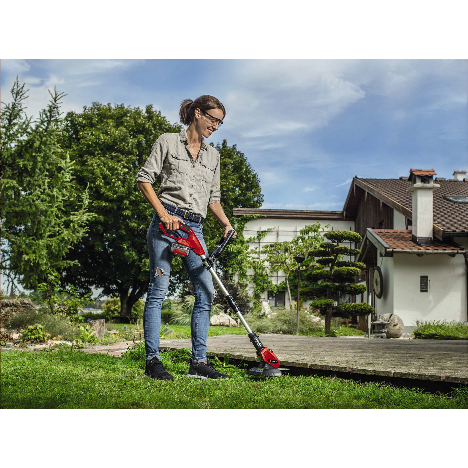Frau verwendet einen Kantenschneider, um Gras um eine Holzterrasse in einem Garten zu trimmen.