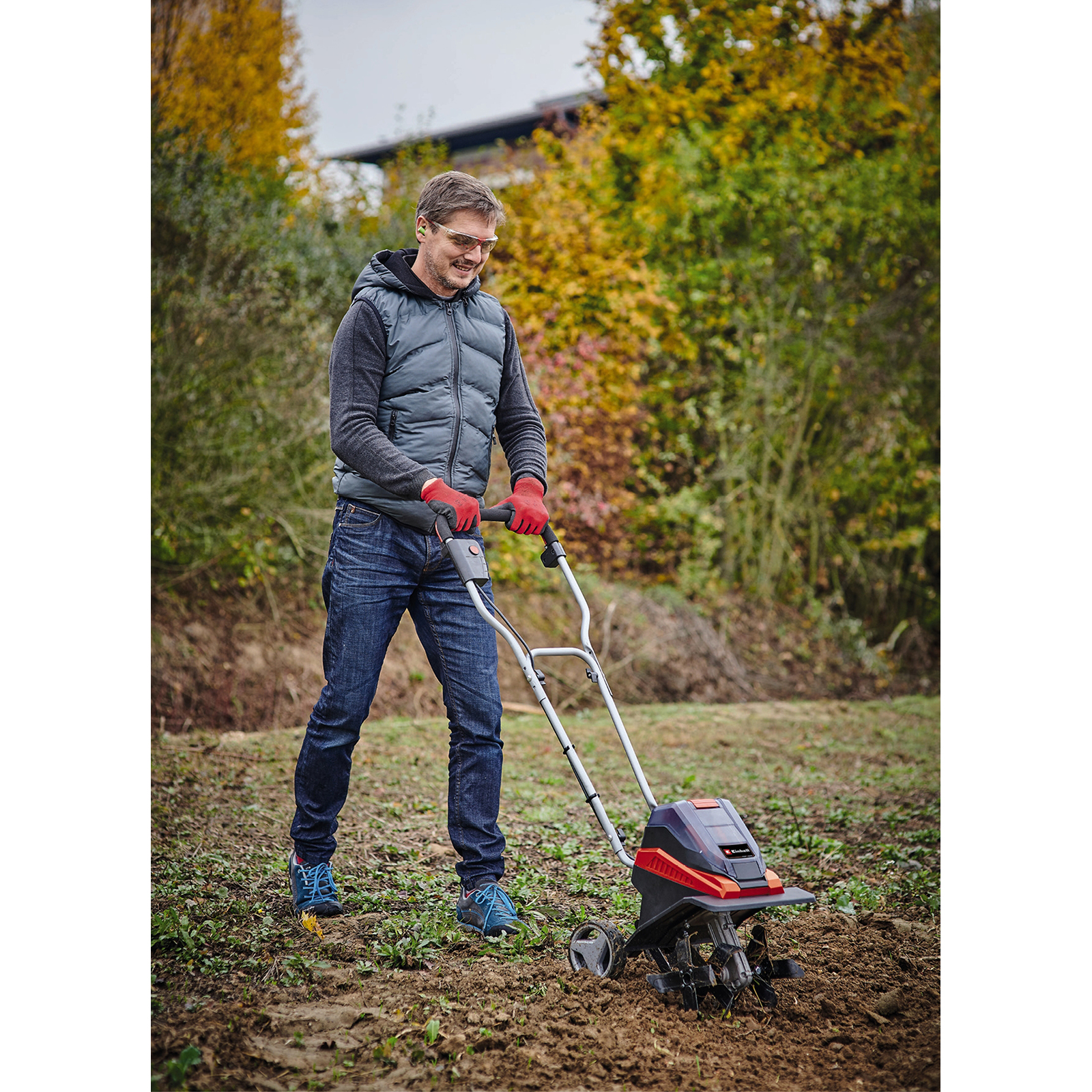 Ein Mann benutzt eine Fräse auf einem Feld. Er trägt eine Jacke, Handschuhe und eine Schutzbrille. Bäume im Hintergrund.