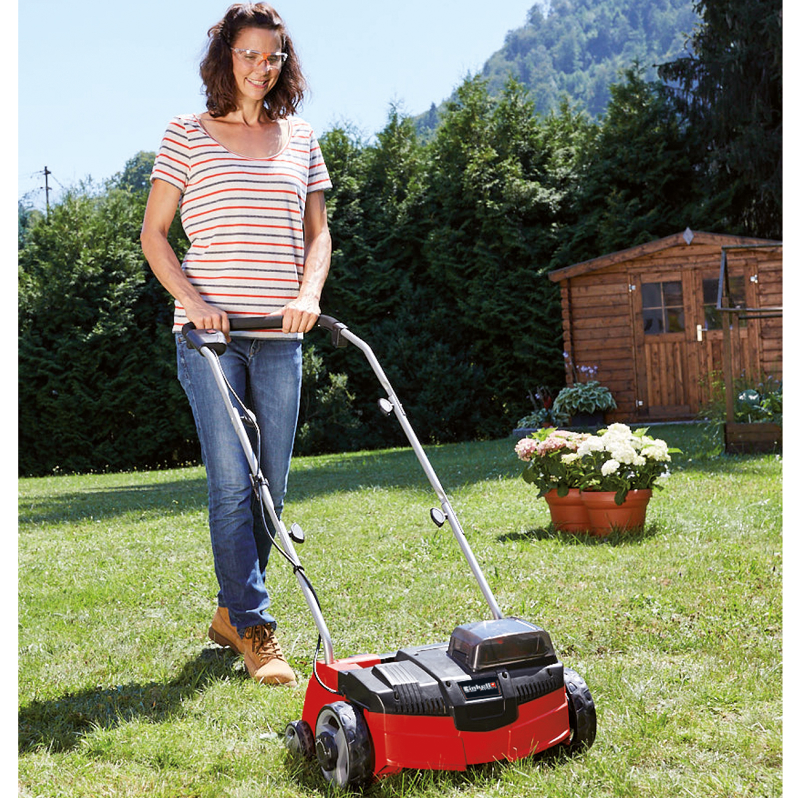 Eine Frau mäht den Rasen mit einem roten Rasenmäher in einem grünen Garten. Ein Holzschuppen und Blumen im Hintergrund.