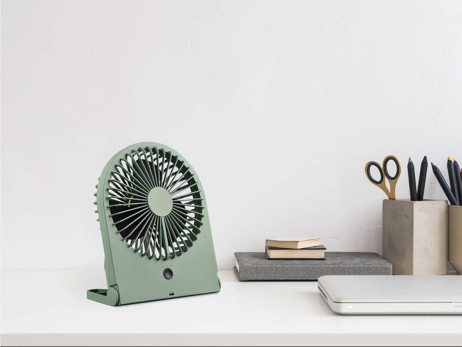 Ein grüner Schreibtischventilator steht mit anderen Gegenständen auf einem weißen Schreibtisch. Der Hintergrund ist eine weiße Wand.