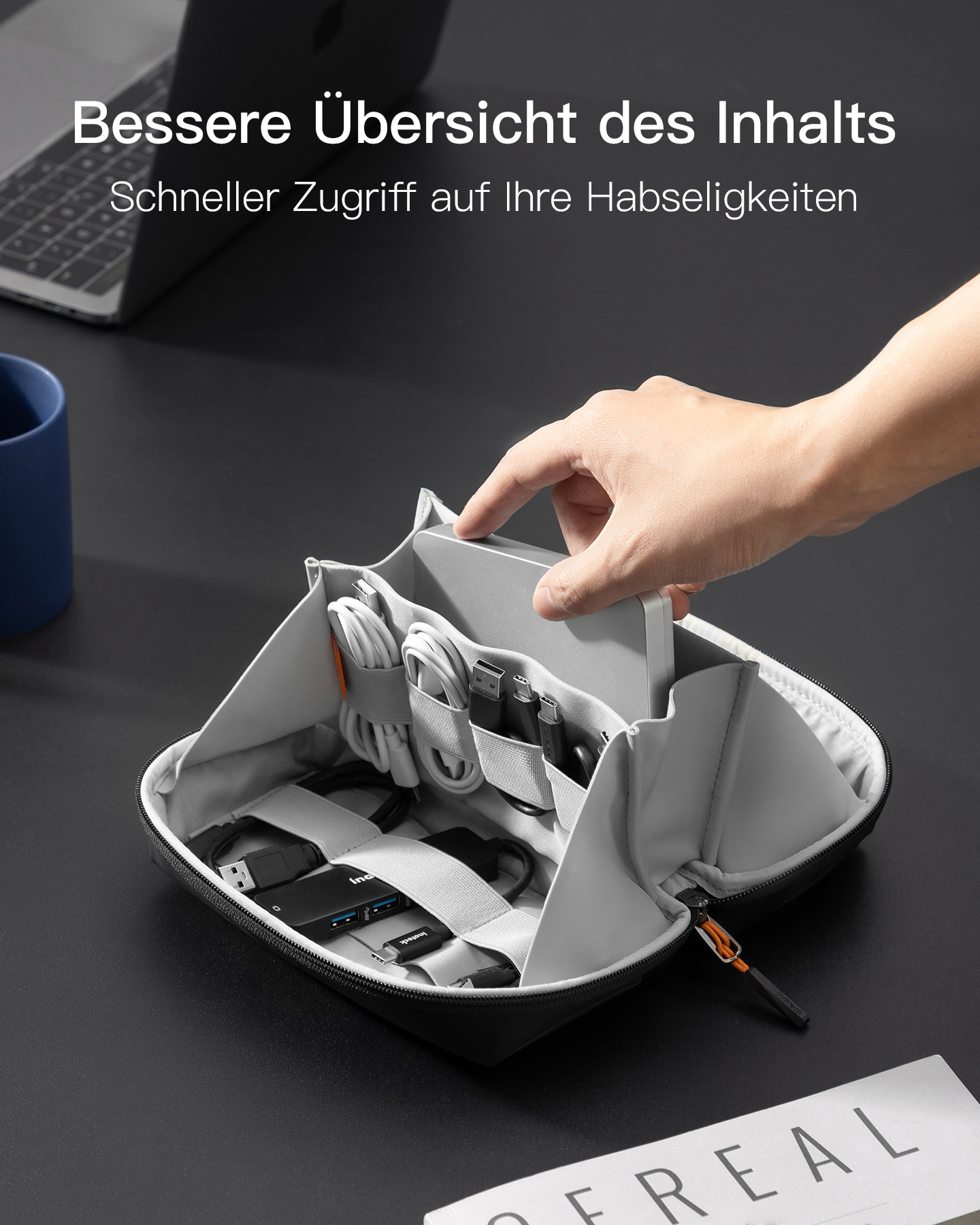 Eine Person greift in eine offene graue Tasche auf einem Schreibtisch, gefüllt mit Kabeln und Elektronik.
