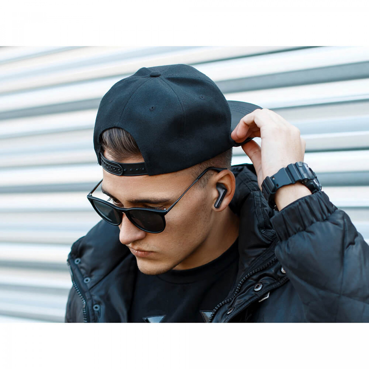 Mężczyzna w okularach przeciwsłonecznych i czapce, trzymający słuchawkę, z miejskim tłem.