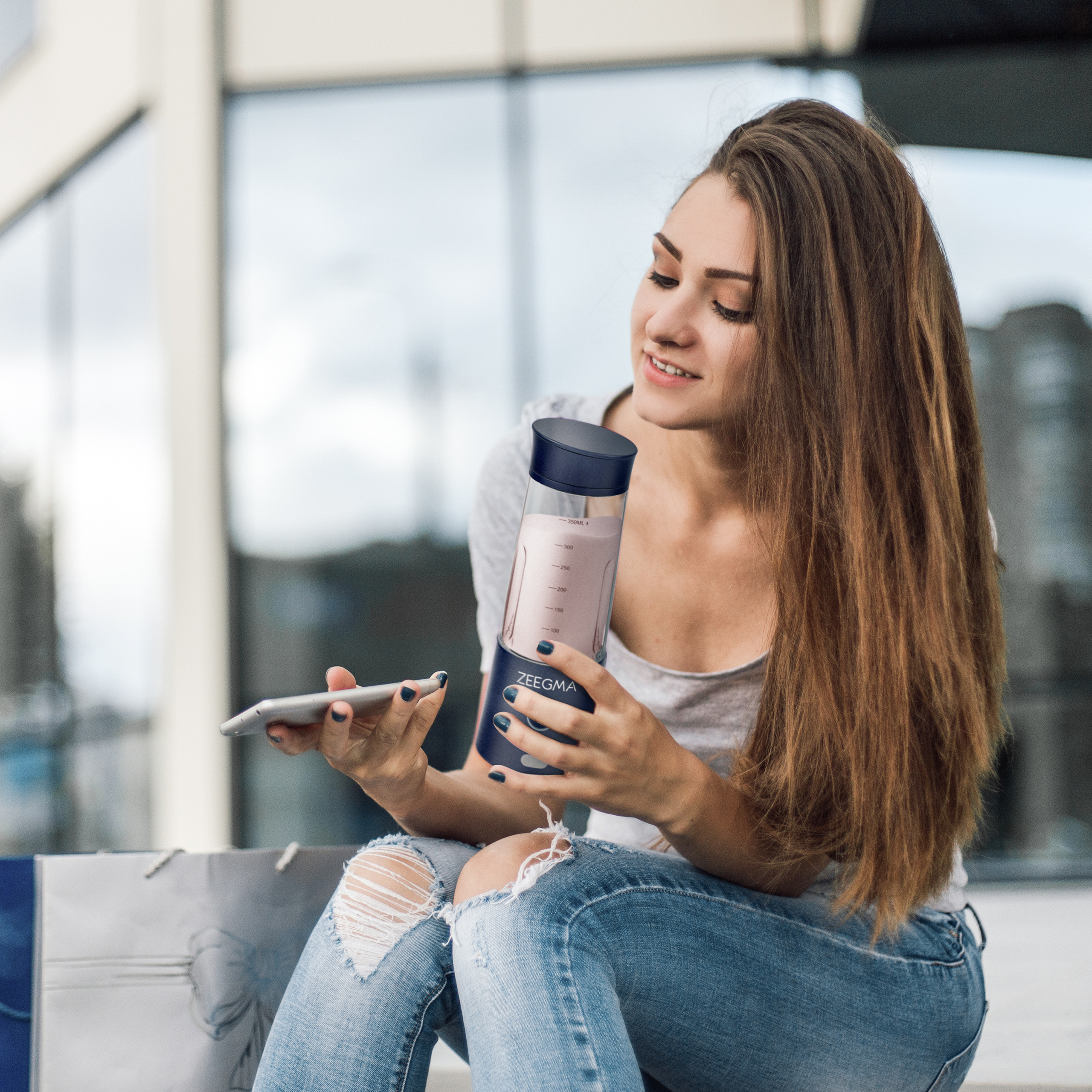 Kobieta z długimi brązowymi włosami trzyma niebieski blender, patrząc na niego. Torby na zakupy na ziemi.