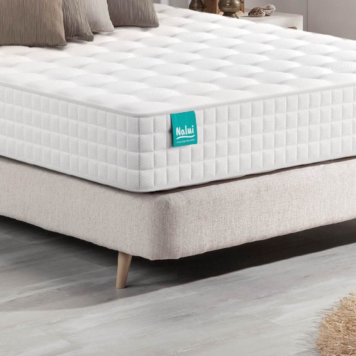 Colchón blanco con etiqueta azul sobre base beige. Almohadas arriba.