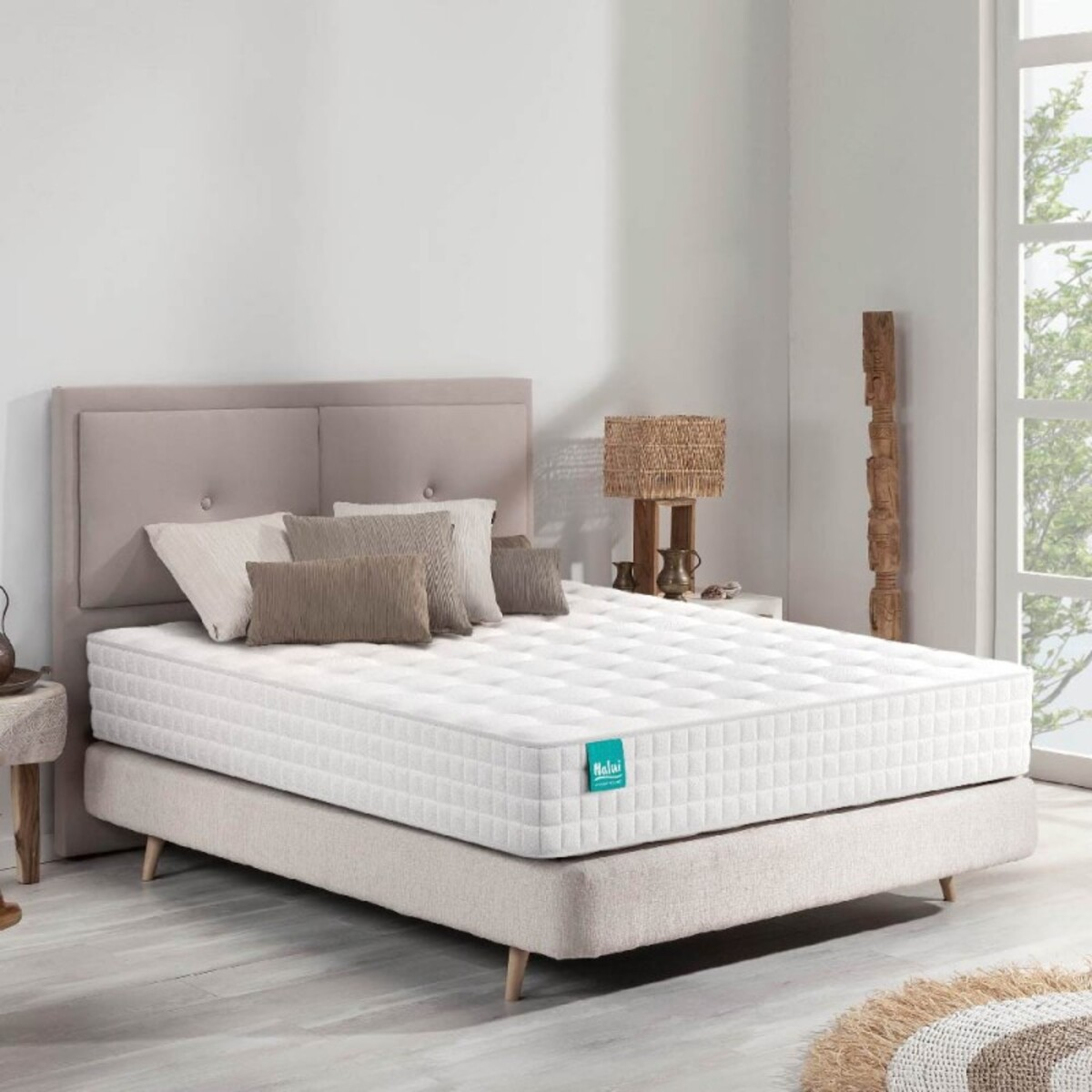 Escena de dormitorio con cama, almohadas y lámpara. La cama tiene un colchón blanco. El fondo es una pared.