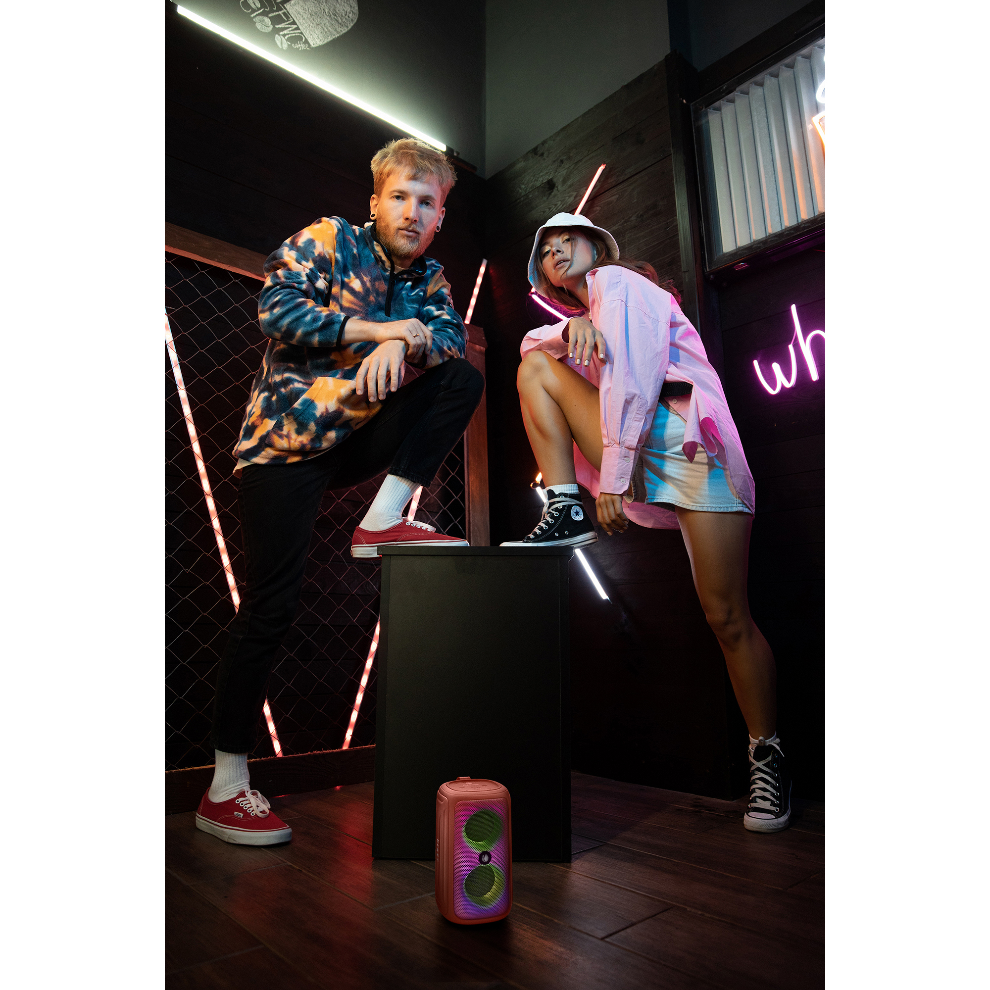 Dwie osoby stojące. Mężczyzna w bluzie z kapturem tie-dye. Kobieta w różowym topie. Głośnik na czarnym stojaku.