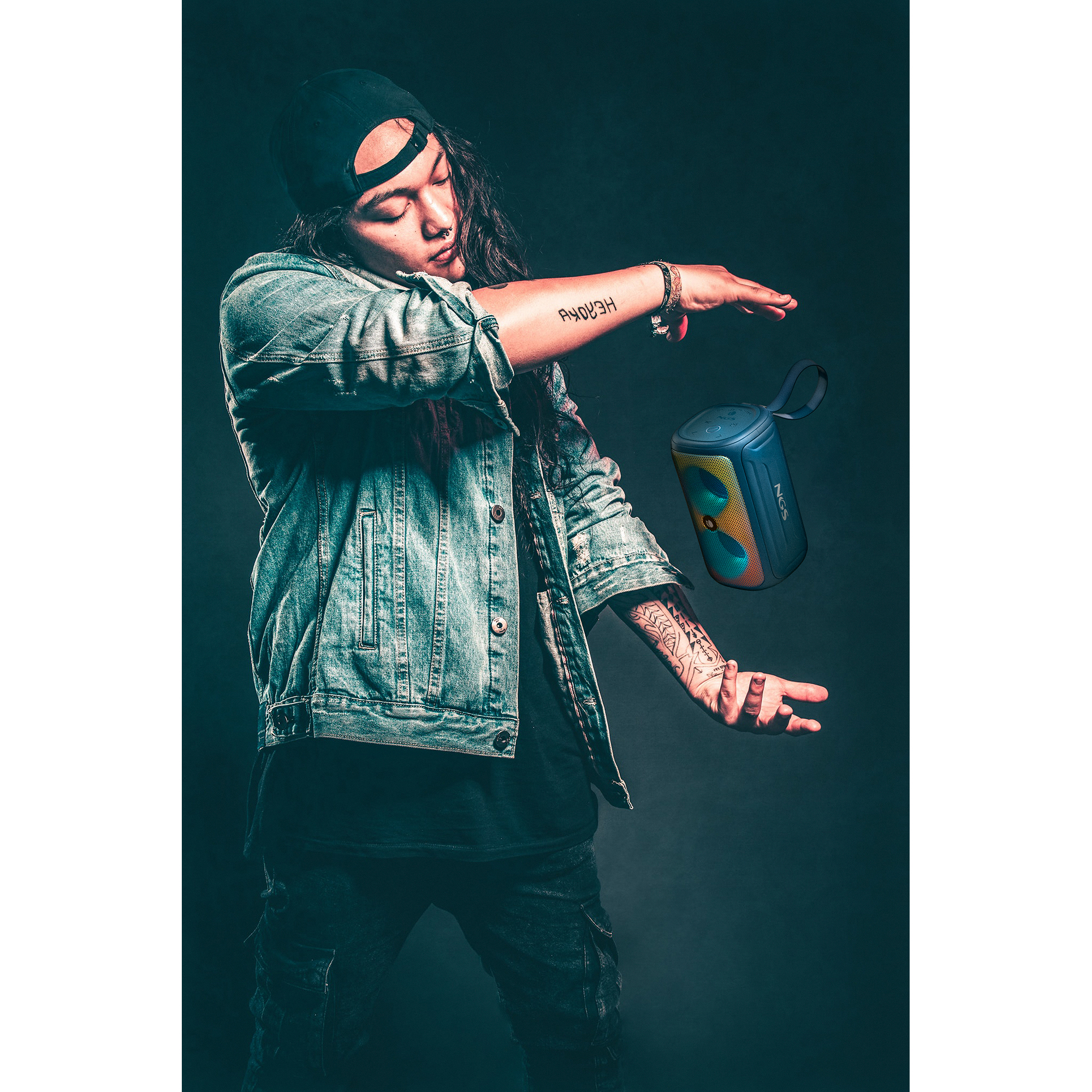 Osoba w dżinsach i dżinsowej kurtce rzuca niebieskim głośnikiem w powietrze na ciemnym tle.