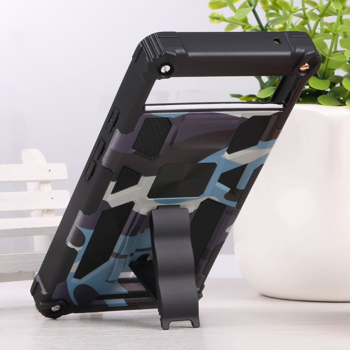 Telefonhülle mit Ständer, Camouflage-Design, abgewinkelt, auf weißer Oberfläche neben Pflanze.