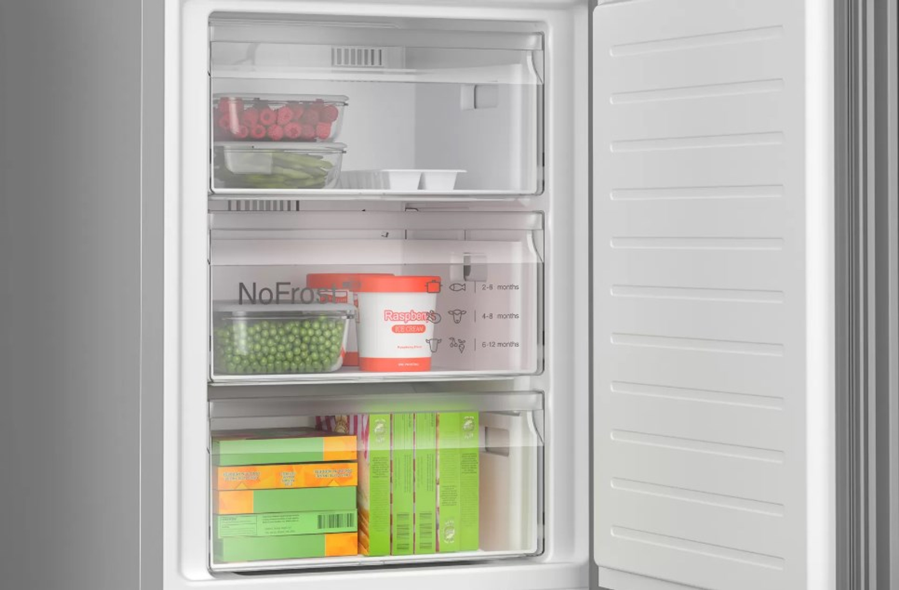 Otwarta lodówka z trzema szufladami wypełnionymi artykułami spożywczymi: malinami, groszkiem i innymi produktami.