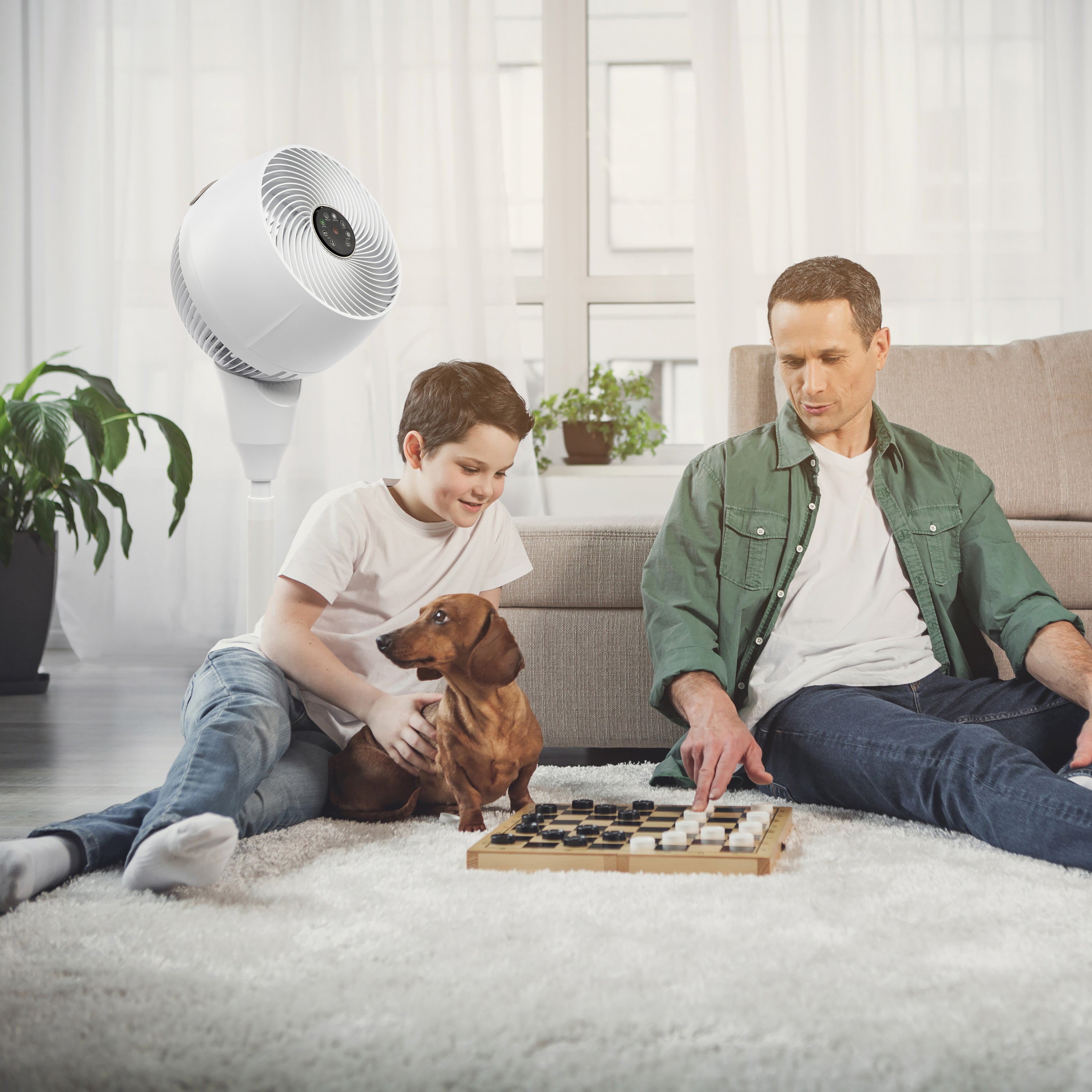 Ojciec i syn grają w warcaby, obok siedzi jamnik. Biały wentylator, biały dywan, kremowa sofa.