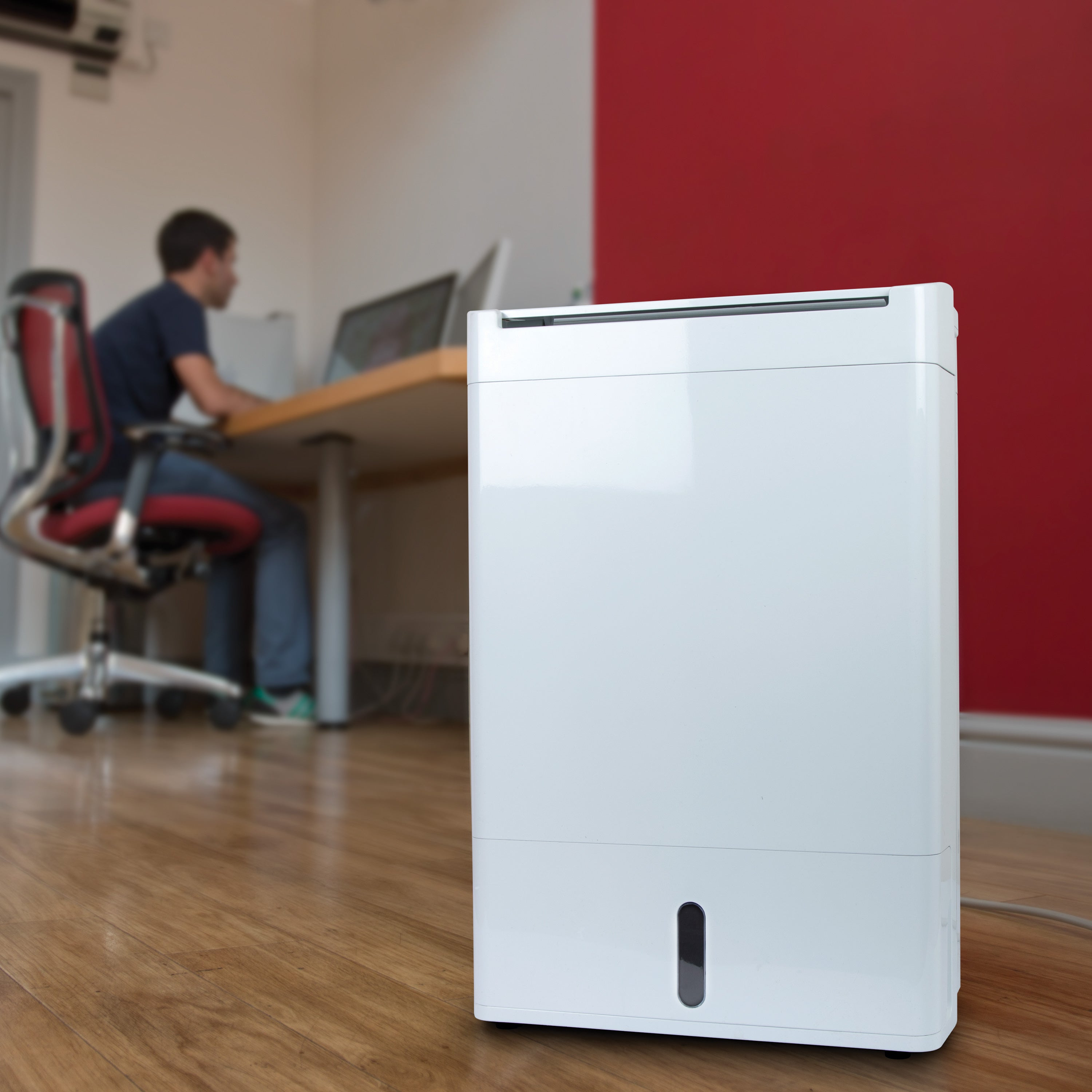Biały osuszacz powietrza na drewnianej podłodze. Osoba w tle pracuje na laptopie.