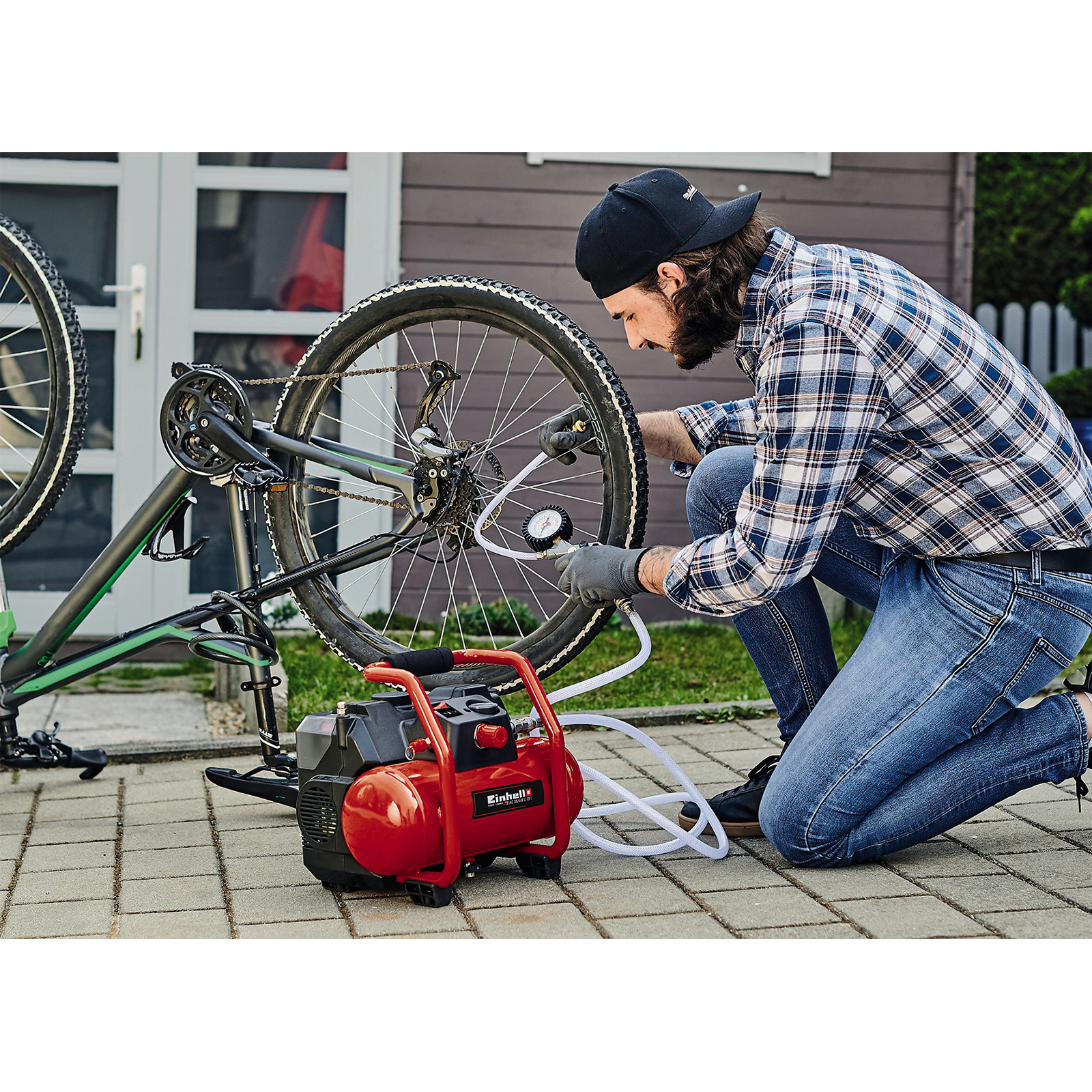 Uomo gonfia pneumatico bici con compressore rosso. Bici parcheggiata, casa dietro.