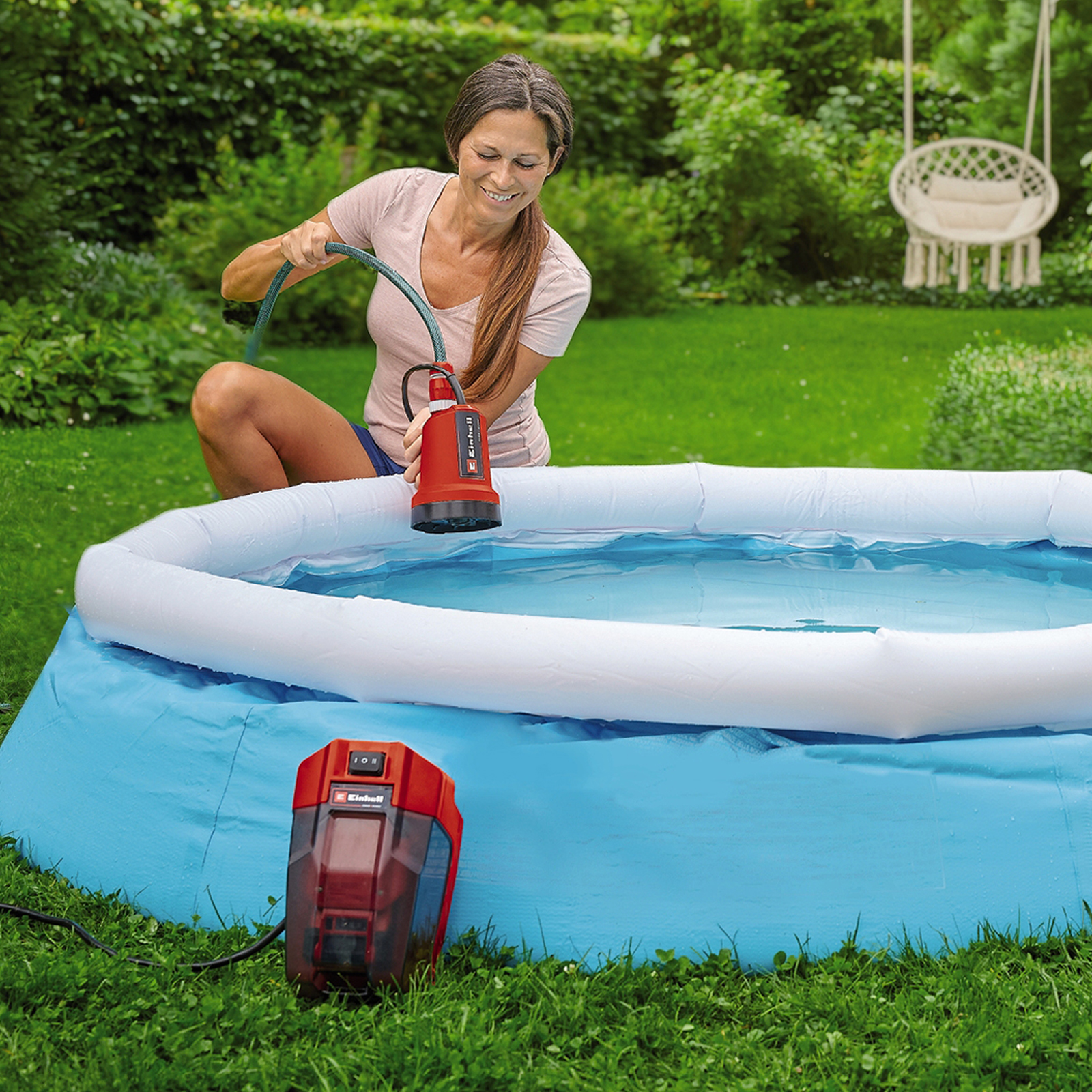 Kobieta używa czerwonej pompy do napełniania niebieskiego basenu w ogrodzie.
