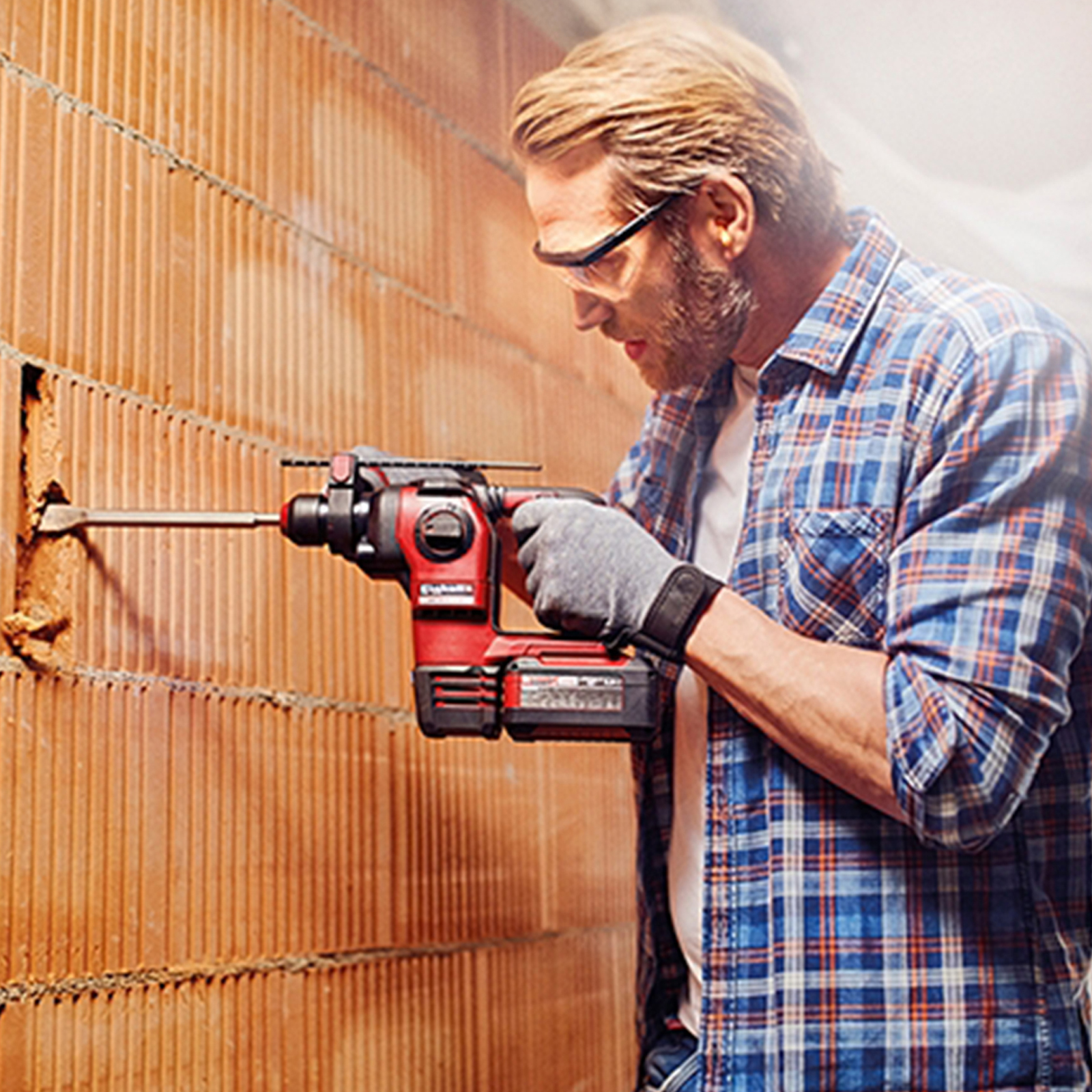 Mężczyzna używa czerwono-czarnej wiertarki na ceglanym murze. Okulary ochronne.