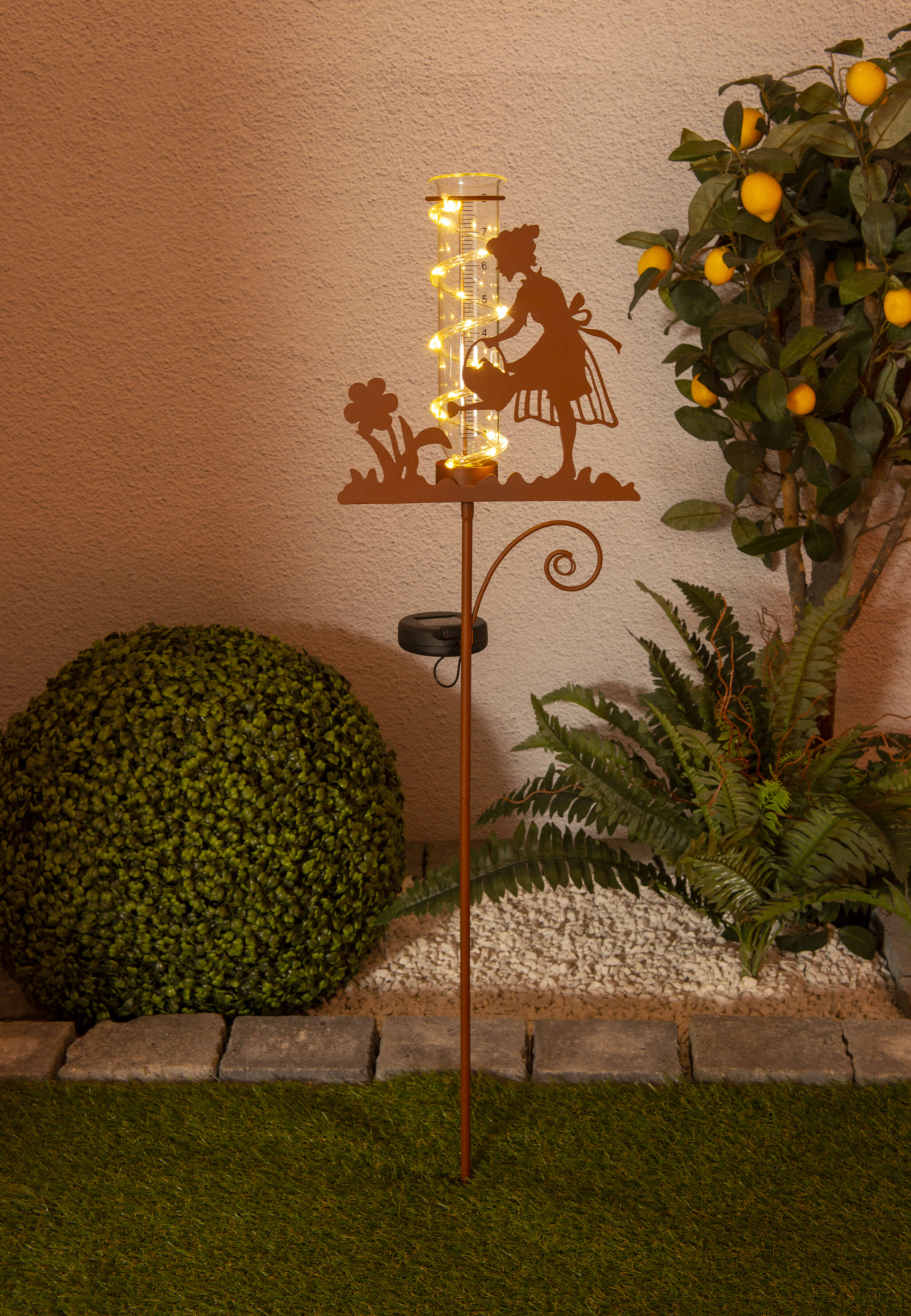 Gartenszene mit einer Metallsilhouette einer Frau, die Blumen gießt. Lichter und grüne Pflanzen sind in der Szene.
