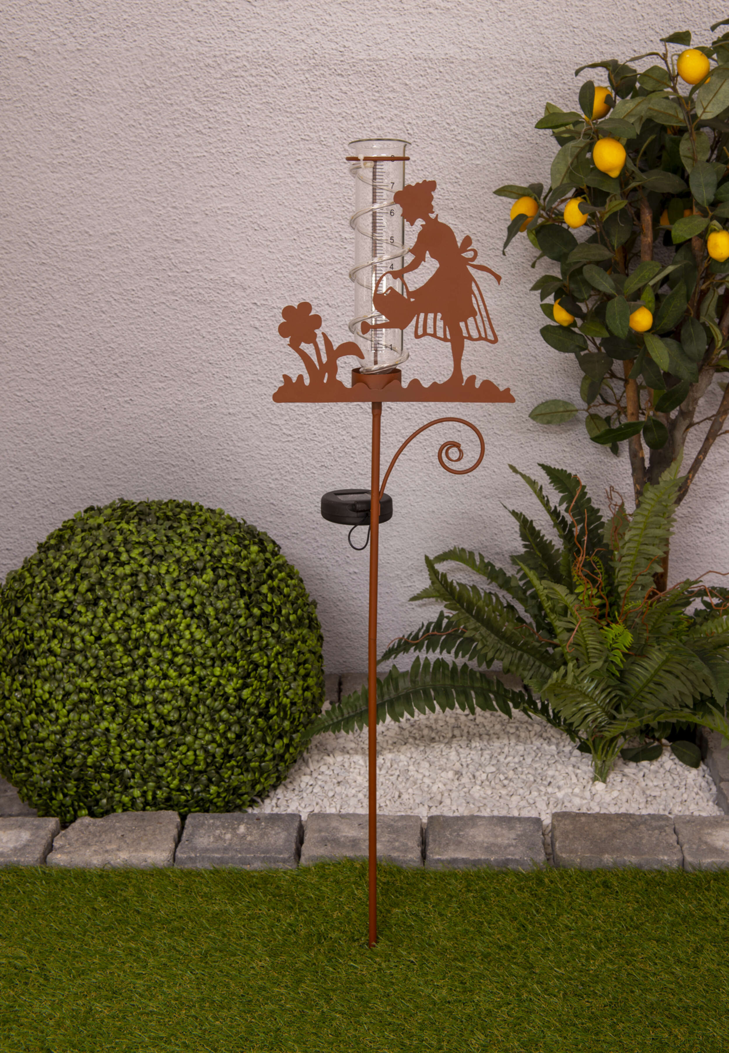 Metall-Regenmesser mit Frauensilhouette, die Blumen gießt, umgeben von Pflanzen und einem Zitronenbaum.