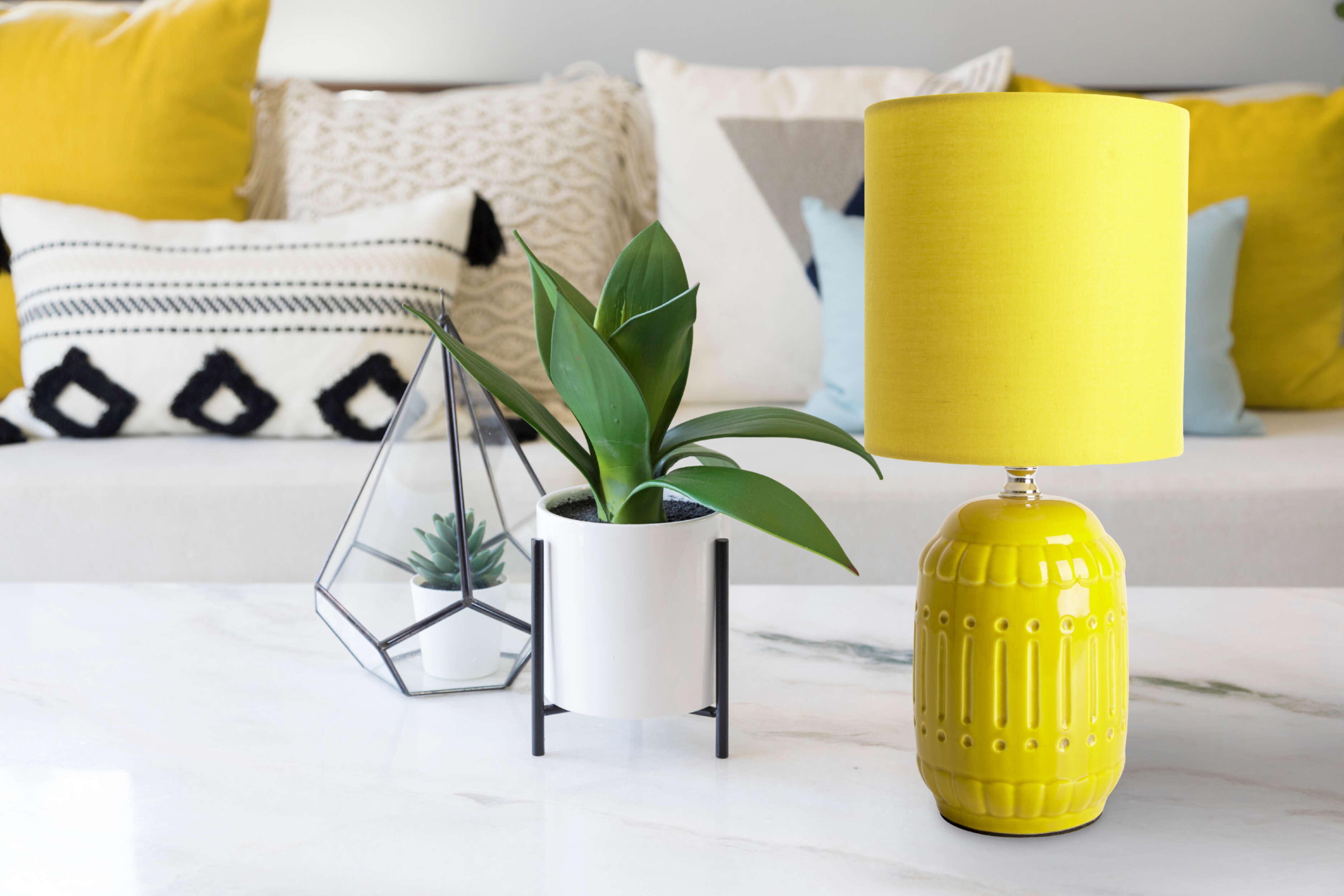 Wohnzimmer mit gelber Lampe, Pflanze und dekorativen Kissen auf weißem Sofa.