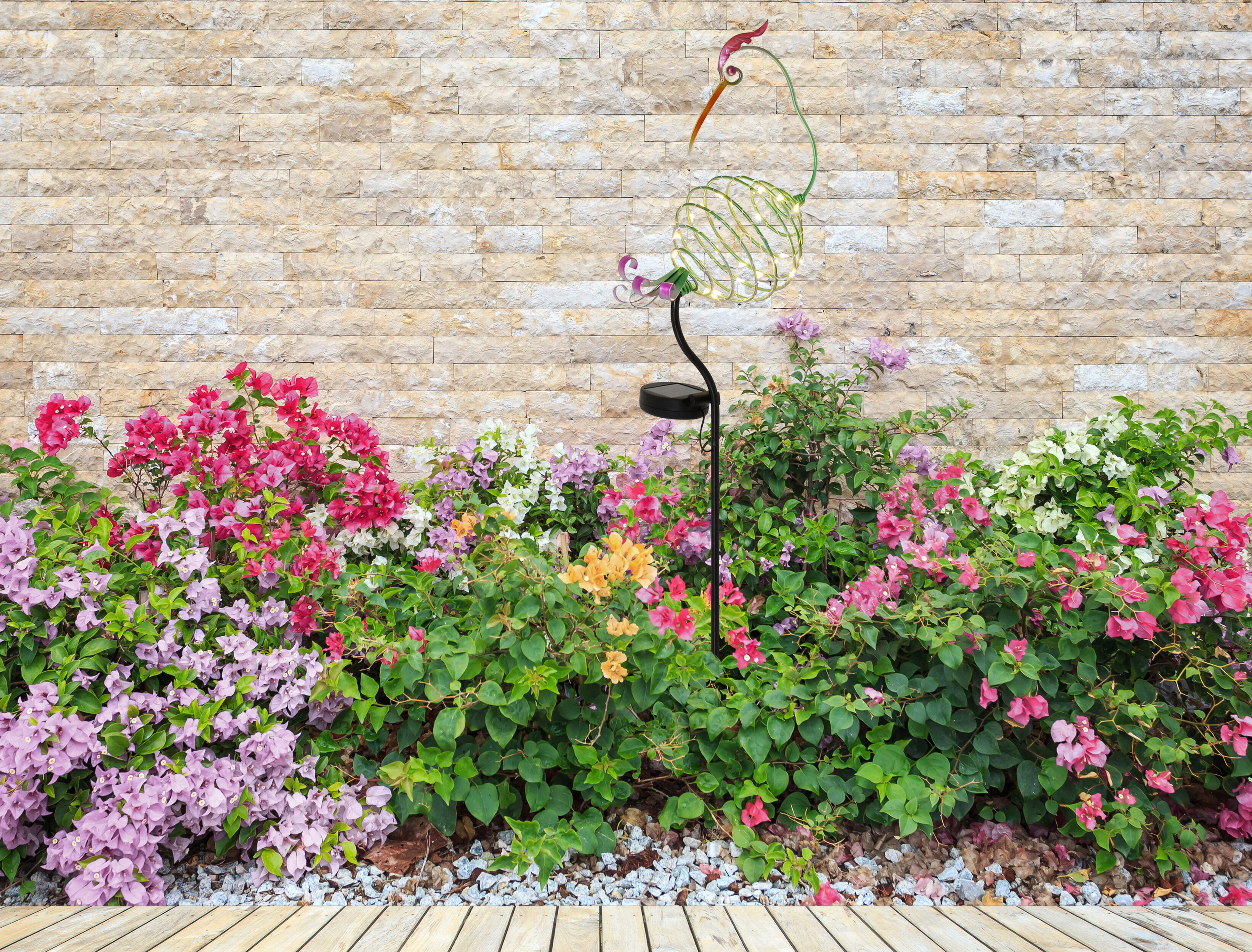 Eine Metallkran-Gartenleuchte vor einer Backsteinmauer und blühenden Büschen.