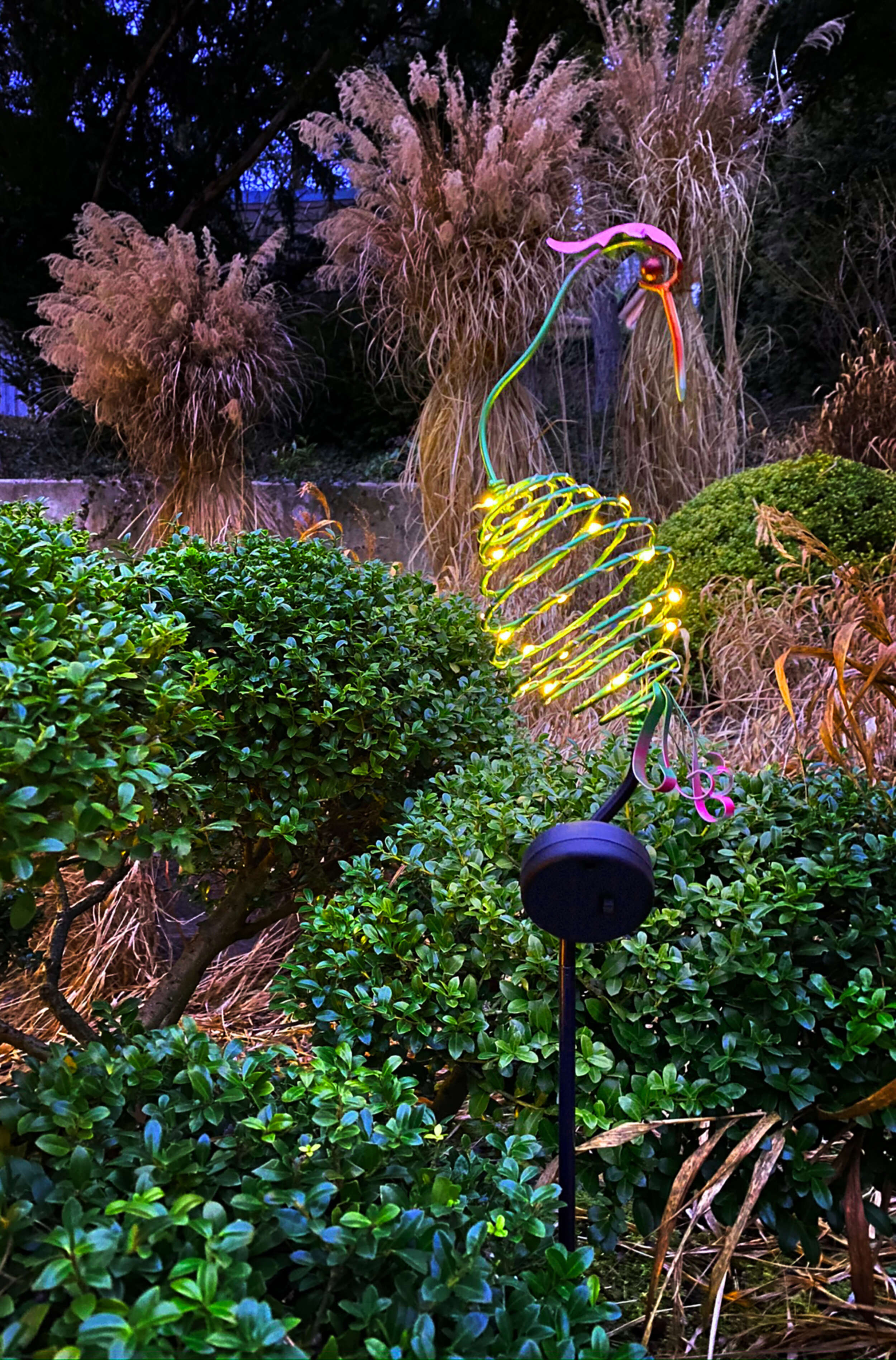 Eine Gartenszene mit einer Kolibri-Pfahllampe, umgeben von grünen Büschen und Pflanzen.
