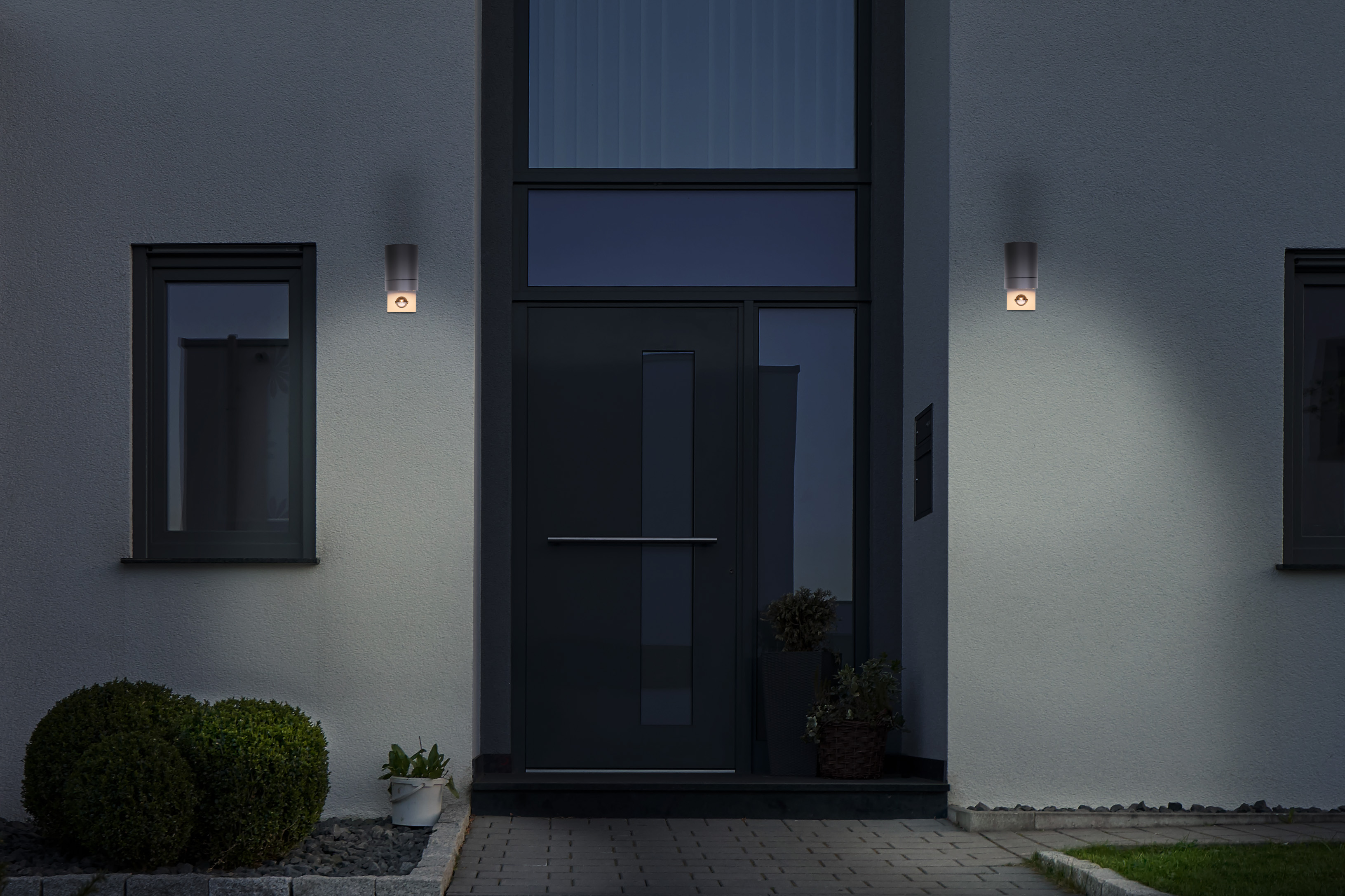 Außenansicht eines Hauses bei Nacht. Zwei Lichter flankieren die Tür. Büsche befinden sich im Vordergrund.